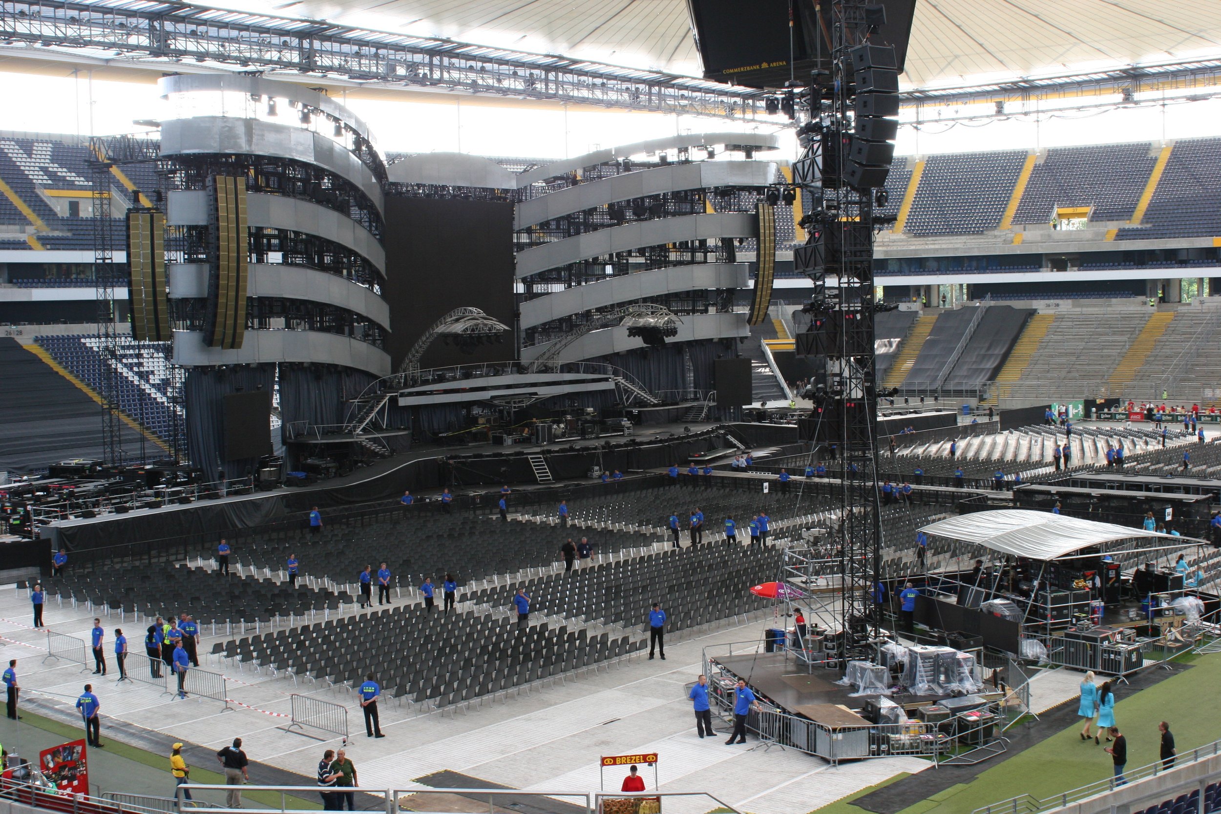 Der Blick auf eine große leere Konzertbühne in einem Stadion, umgeben von viele Stühlen, auf die Bühne sind technische Geräte und Bühnenstrukturen aufgebaut, im Vordergrund sind Security-Personal und einige Besucher zu sehen.