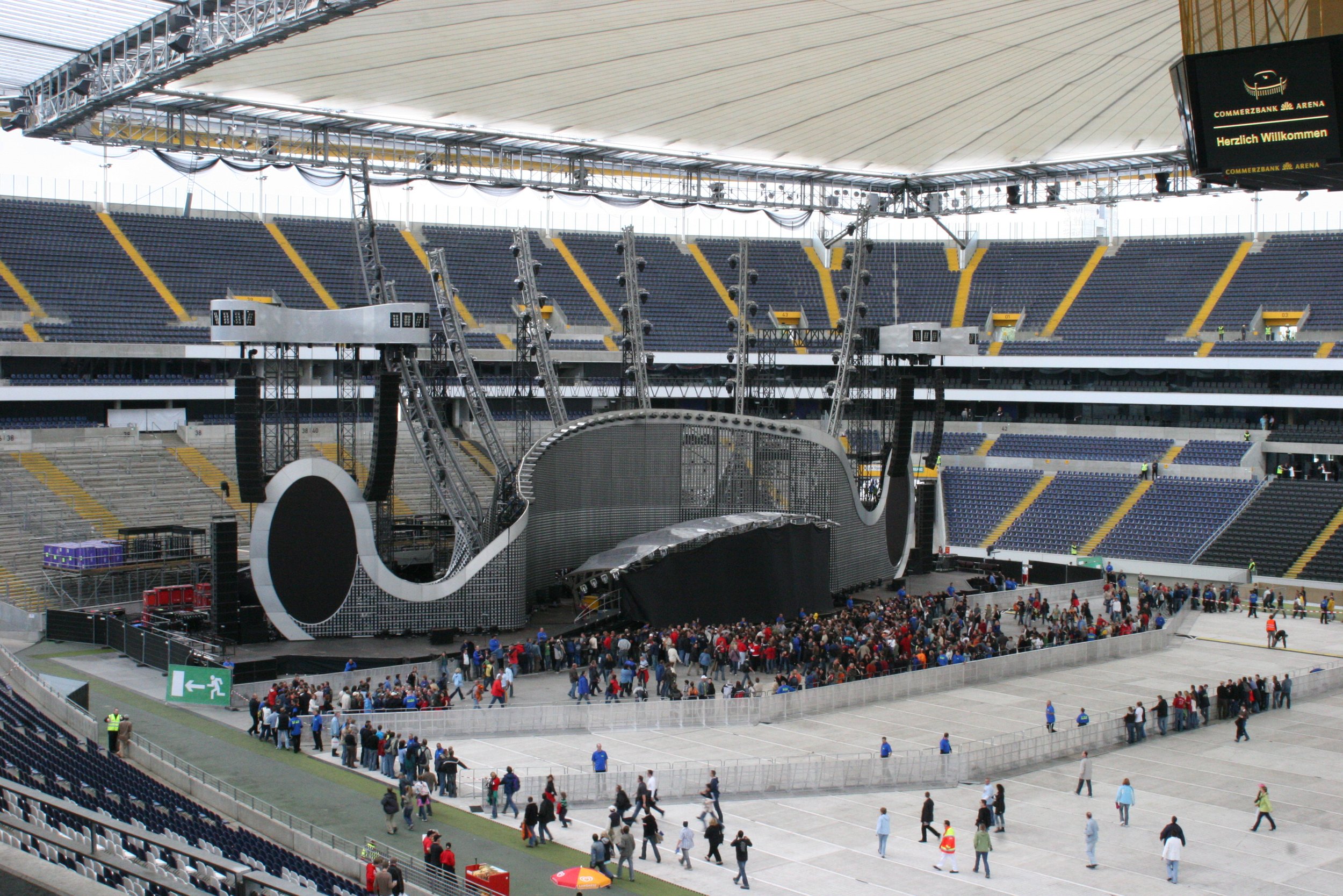 Innenansicht eines großen Stadions mit einer Großbildleinwand und einer absperrbaren Bühne im Zentrum. Es sind viele Menschen unterwegs, vermutlich für eine Veranstaltung im Aufbau.