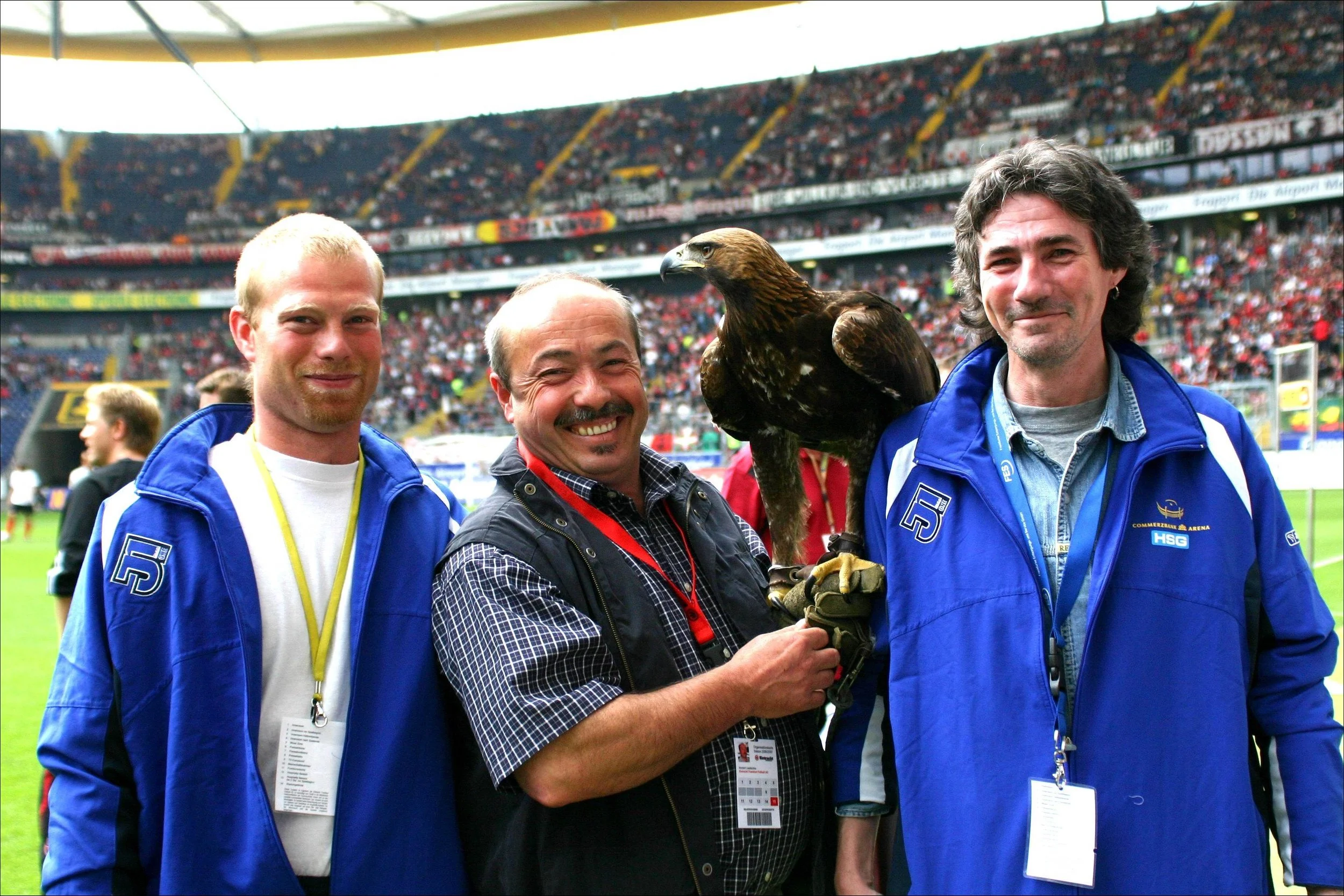 Drei Männer, einer mit Greifvogel auf der Hand, im Stadion mit Zuschauern im Hintergrund.