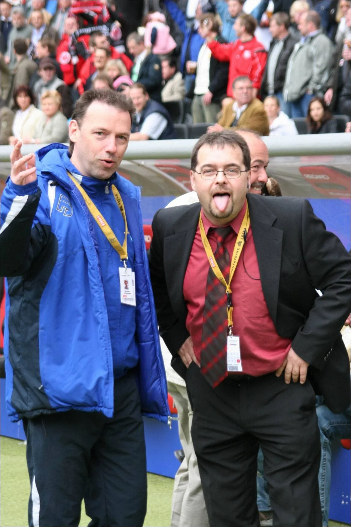 Zwei Männer bei einem Fußballspiel, eines trägt eine blaue Jacke, das andere trägt einen schwarzen Anzug mit rotem Hemd und Krawatte, stehen vor einer Menschenmenge im Stadion. Der Mann im Anzug streckt die Zunge heraus.