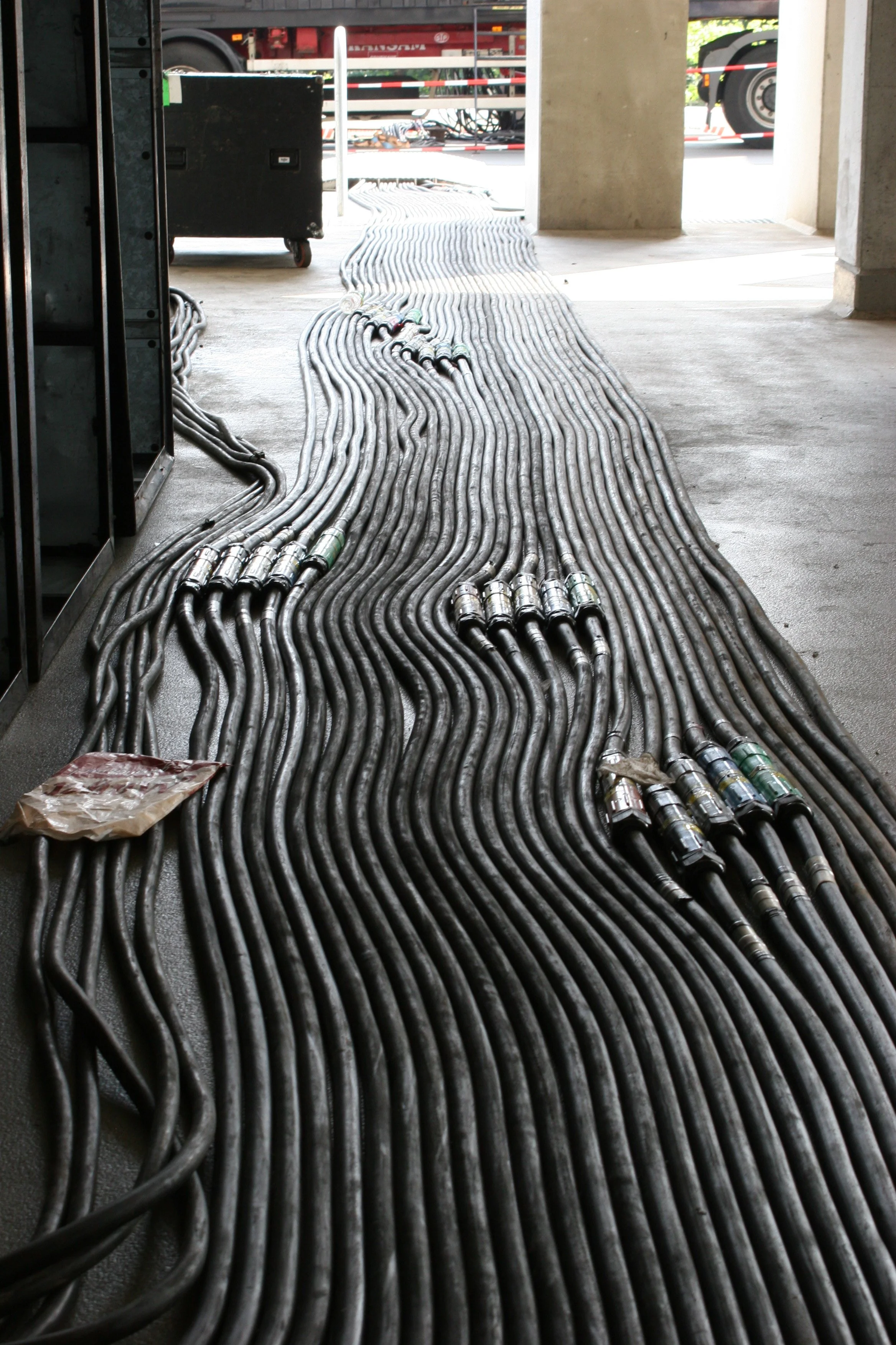 Mehrere schwarze Kabel verlaufen auf dem Boden in einem Gebäude, möglicherweise im Bereich einer Baustelle oder eines technischen Raums, mit einem offenen Eingang und einem Transportfahrzeug im Hintergrund.