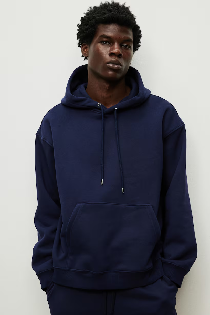 A young man with dark skin and curly hair wearing a navy blue hoodie and matching sweatpants, standing against a plain white background.