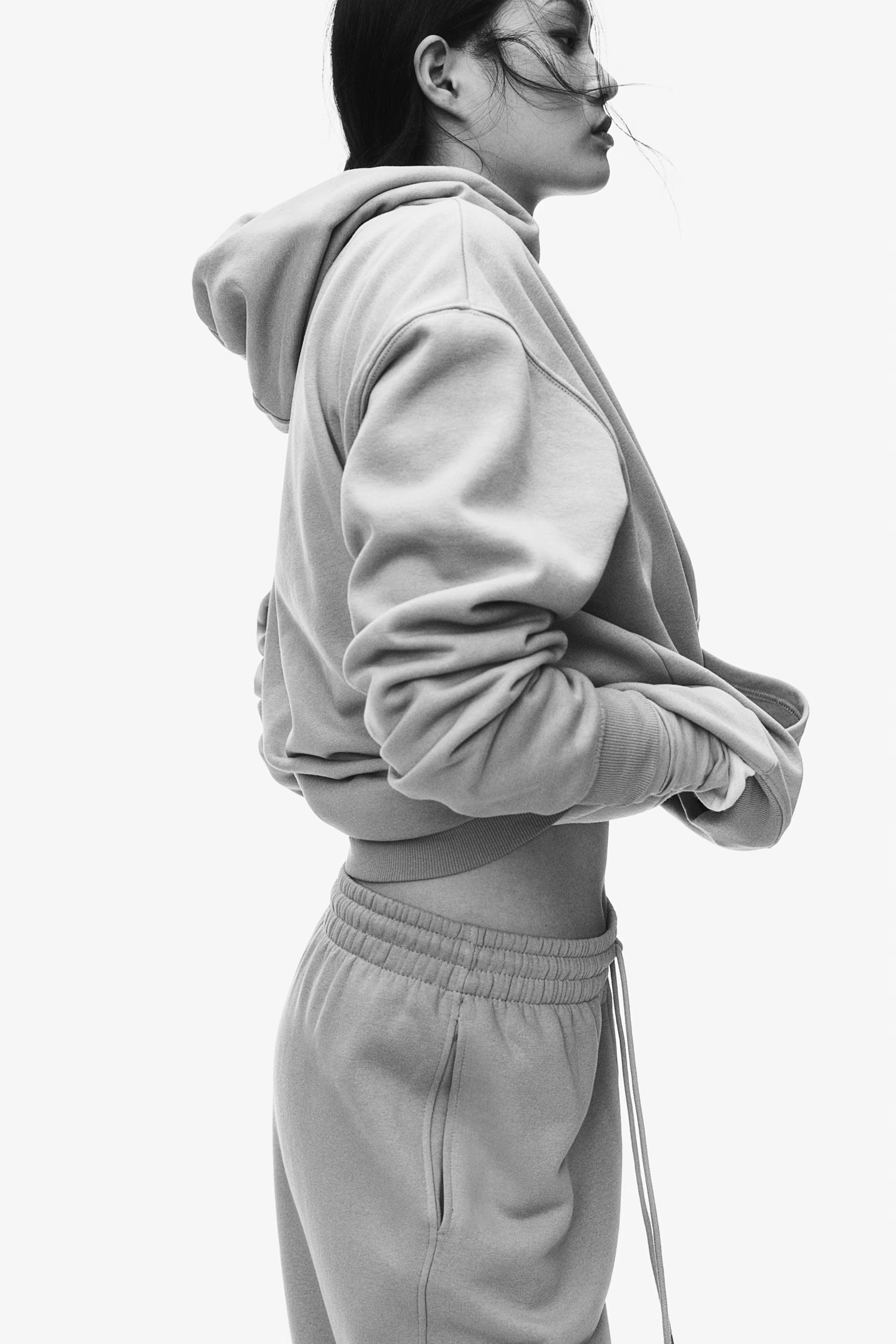 A side view of a woman in a hoodie and sweatpants, with her hands in her pockets, against a plain background.