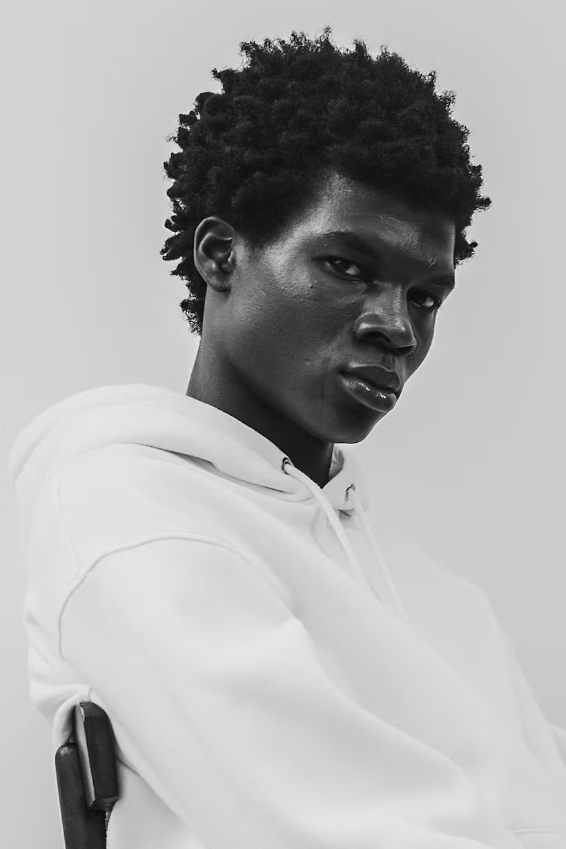 A black and white portrait of a young man with natural, textured hair, wearing a light-colored hoodie, looking intently at the camera with a serious expression.