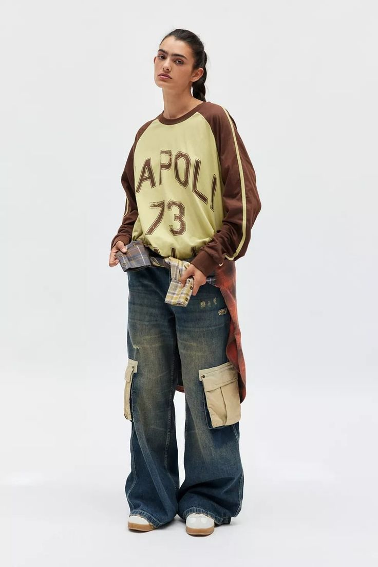 A young woman with dark hair in a ponytail, wearing a vintage-style oversized sweatshirt with 'APOL 73' printed on it, baggy jeans with cargo pockets, and brown and white sneakers, standing against a plain white background.