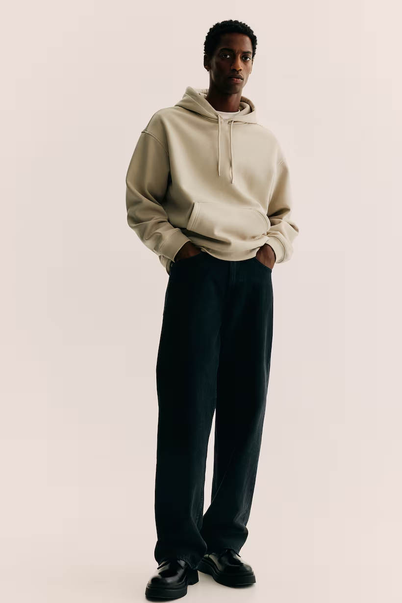 A young man standing with his hands in his pockets, wearing a beige hoodie, black wide-leg pants, and black shoes, against a plain light background.