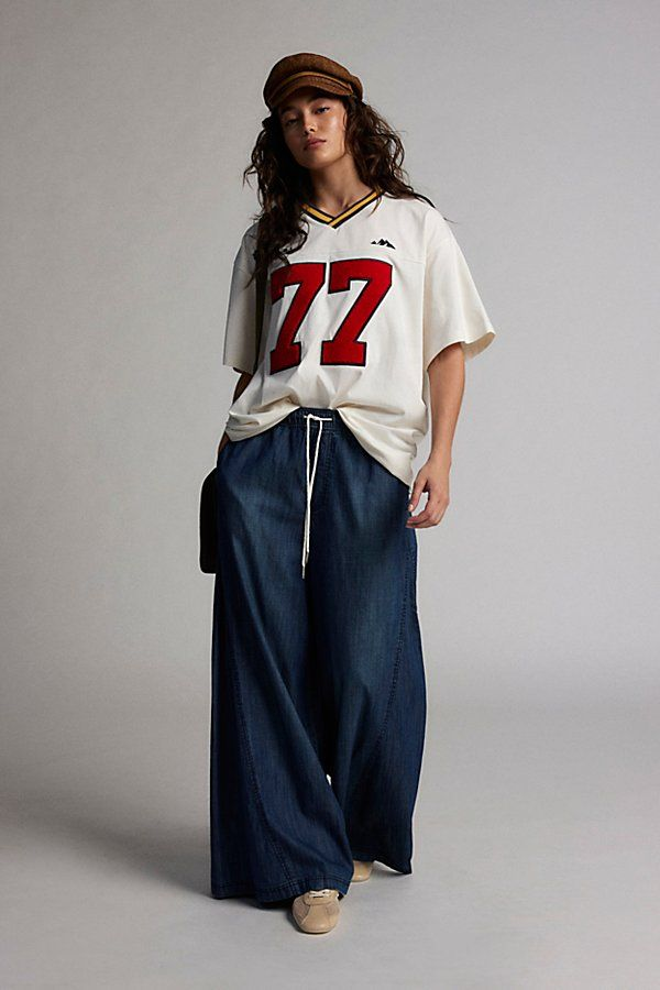 Young woman wearing a beige baseball cap, a loose white sports jersey with the number 77 in red, navy wide-leg pants with a white drawstring, and beige shoes, standing against a plain gray background.