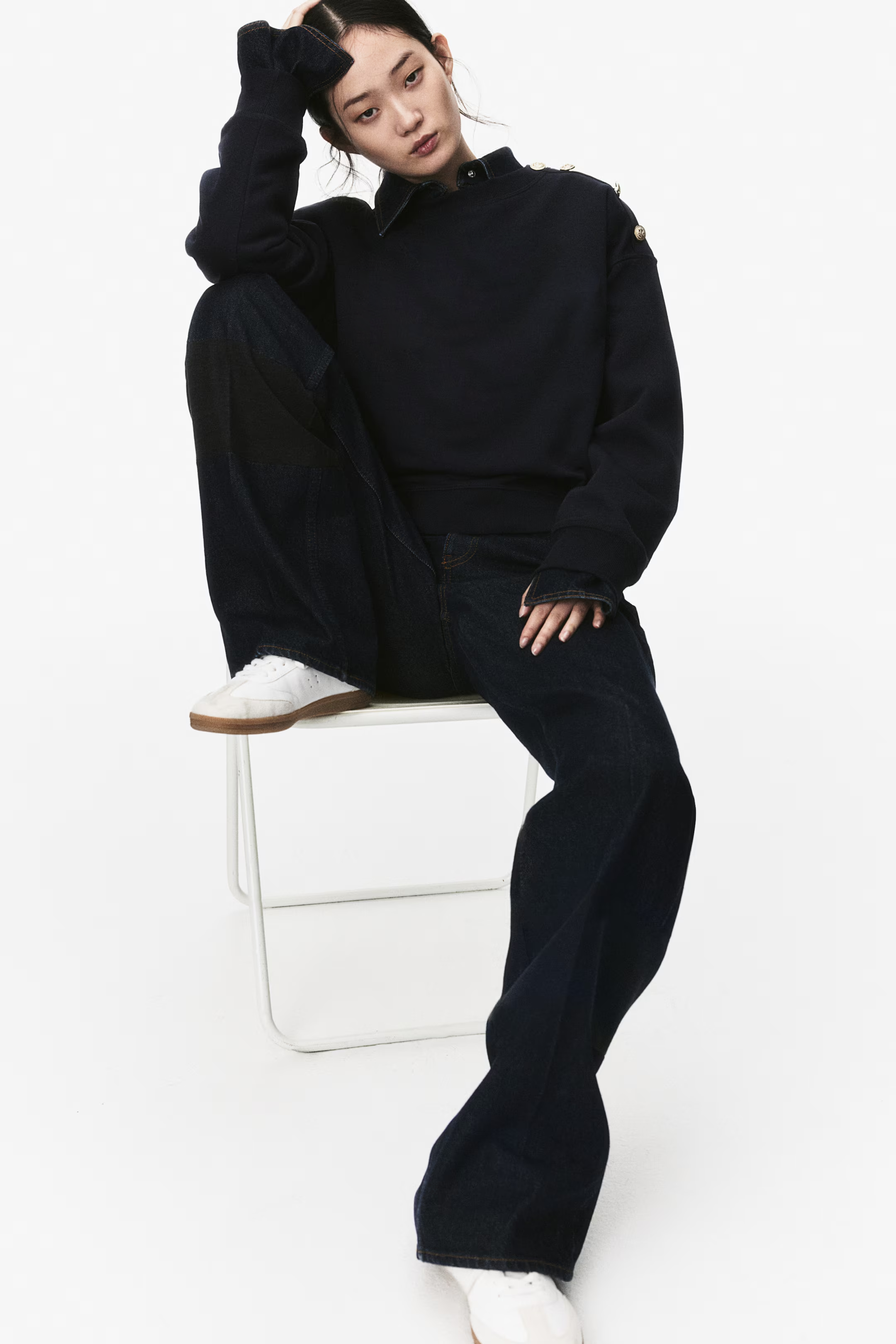A young woman with black hair in a casual black sweatshirt and jeans, sitting on a white chair with one leg resting on the chair, against a plain white background.