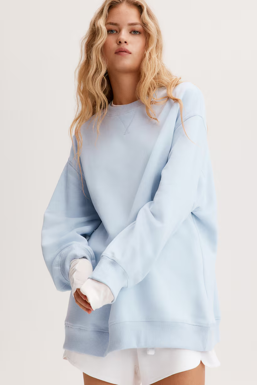 A young woman with long, wavy blonde hair wearing a light blue oversized sweatshirt and white shorts, standing against a plain light background.
