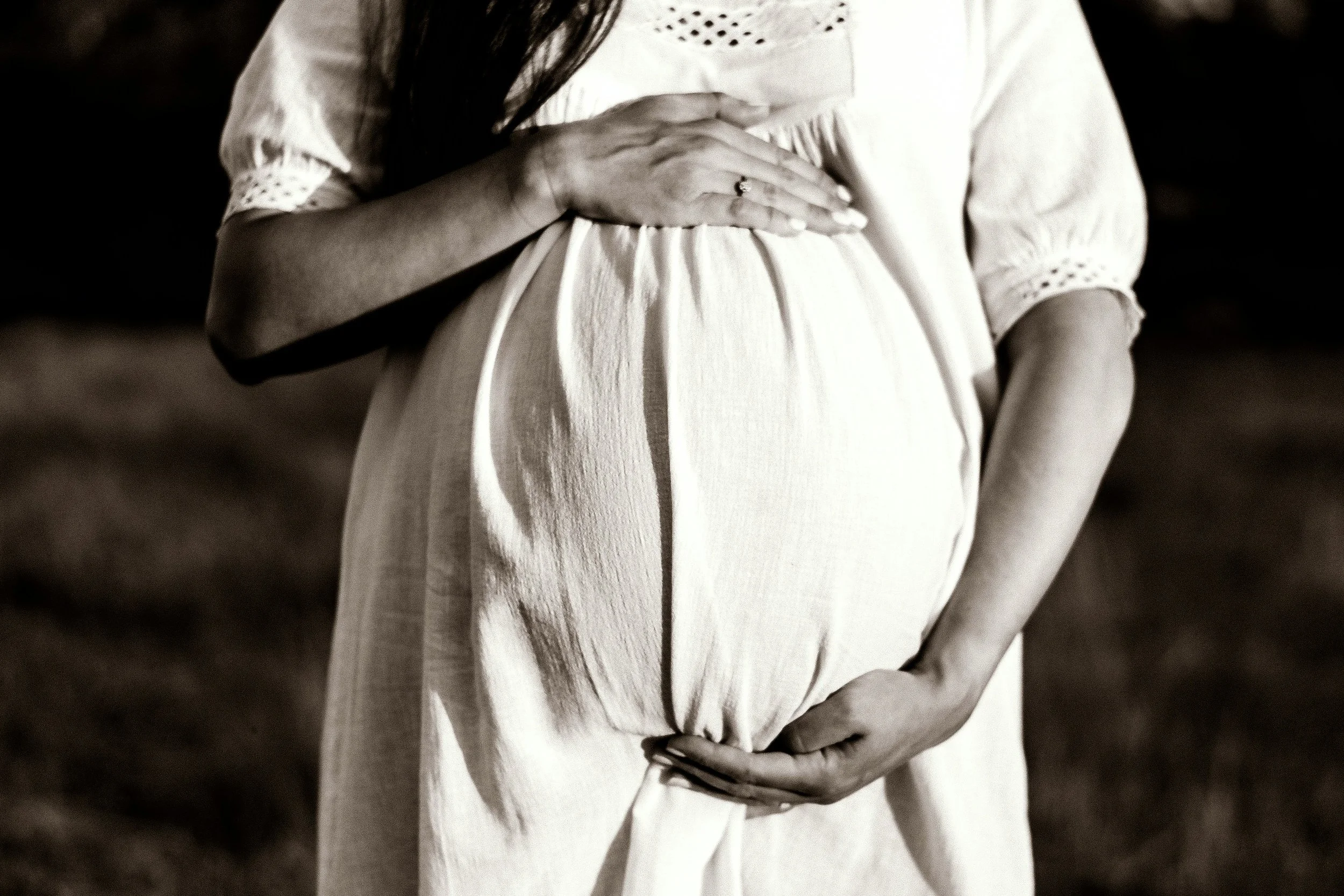 A pregnant woman gently holding her belly with both hands, wearing a light-colored dress, in a natural outdoor setting.