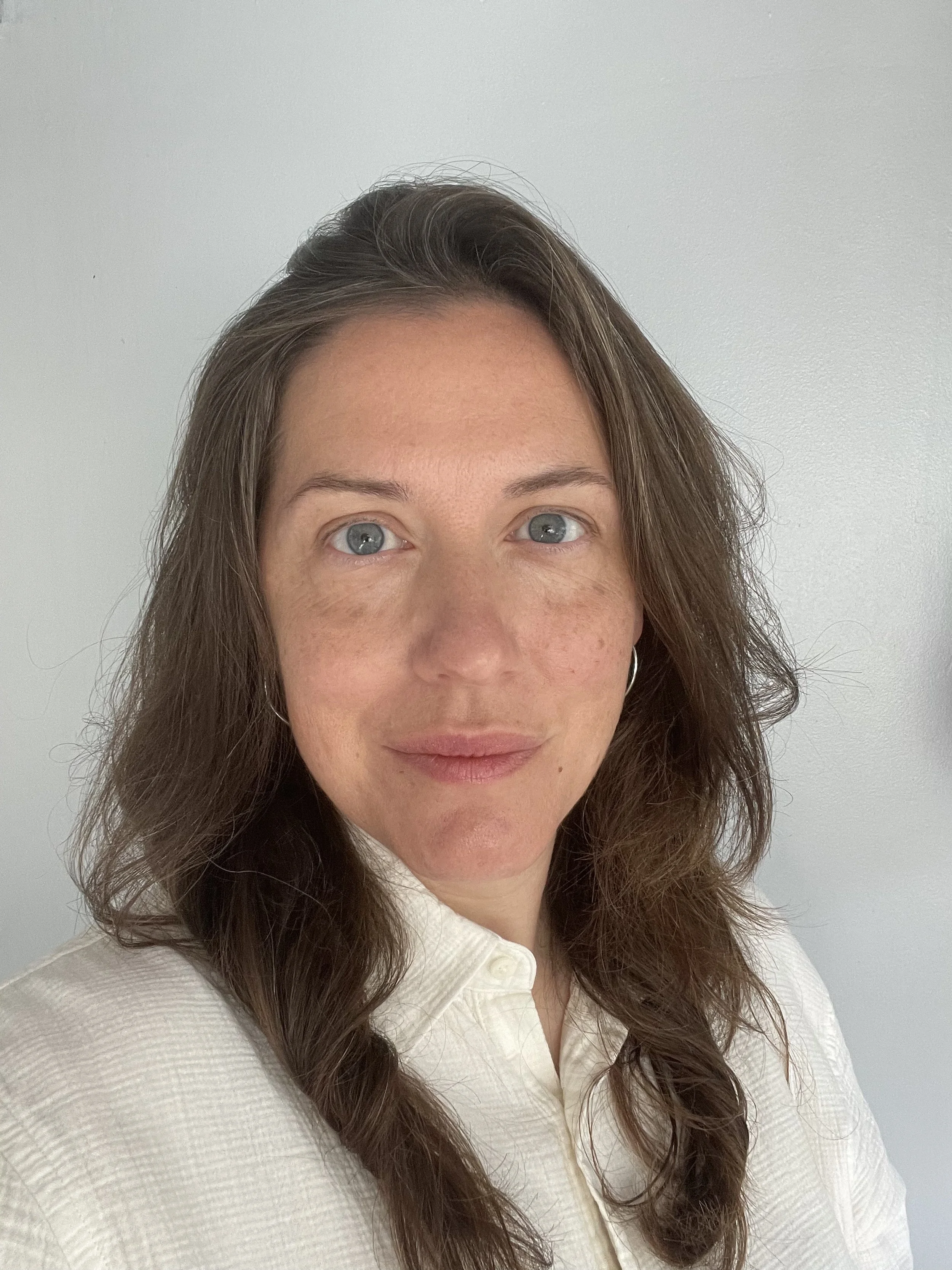 A woman with wavy brown hair and blue eyes, wearing a white shirt, standing against a plain light gray background.