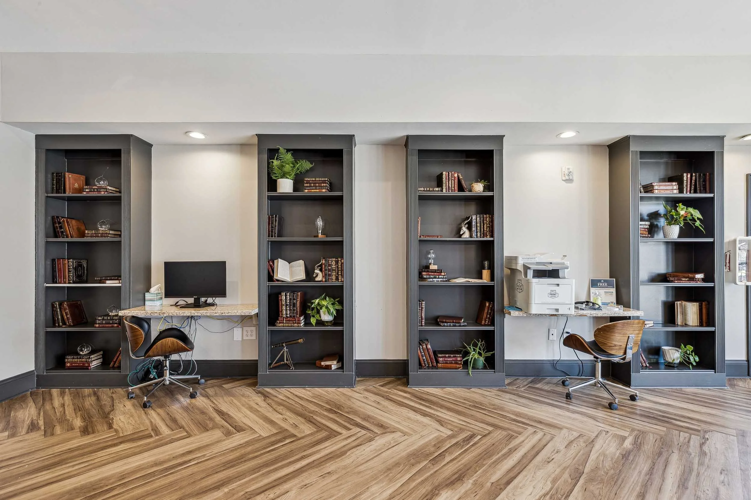 Resident lounge with four black bookshelves filled with books, decorative items, and plants, a desk with a computer and office chair, and a wood floor.