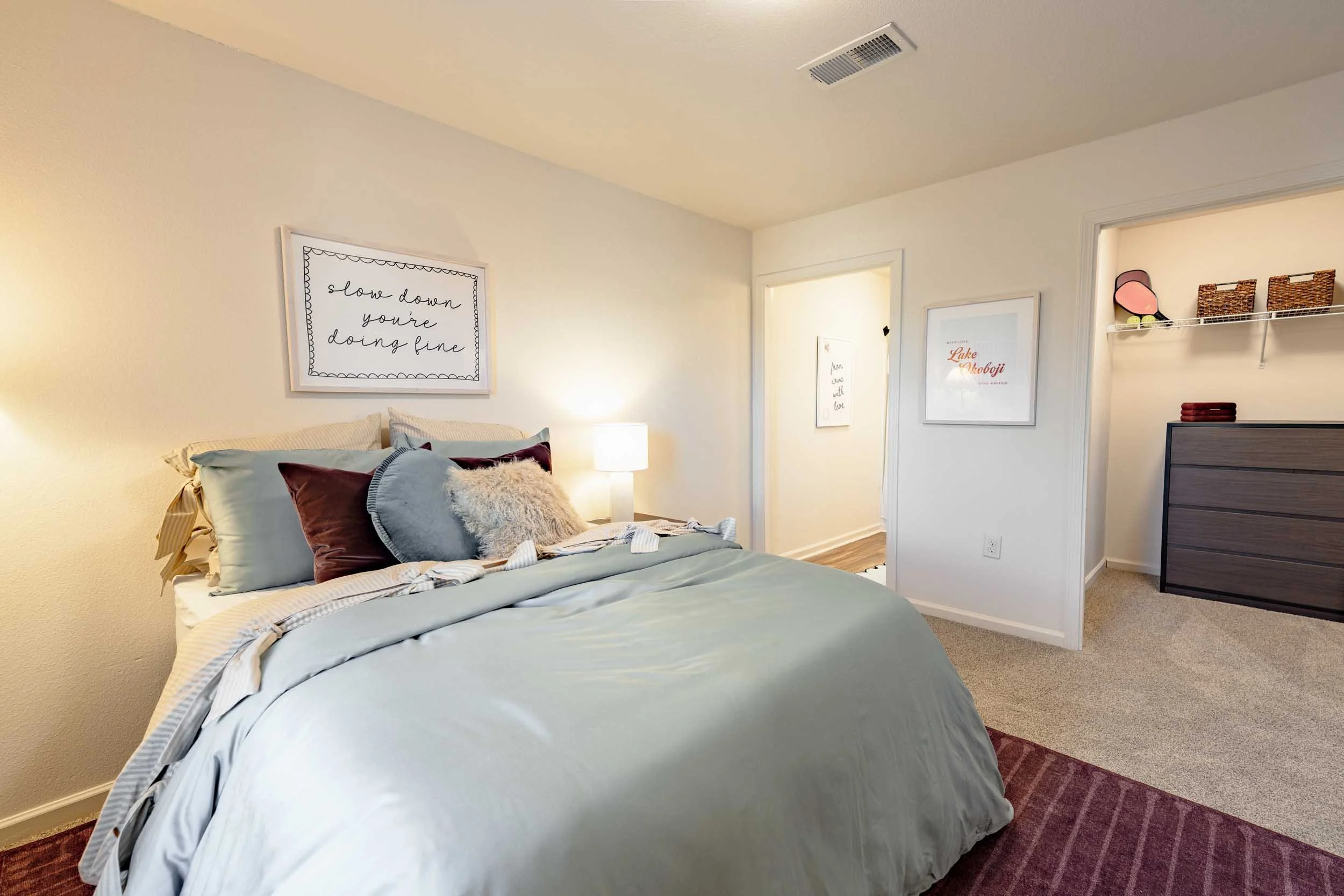 A neatly made bed with light blue sheets and multiple pillows, located in a bedroom with white walls decorated with framed artwork. There is a bedside lamp shining beside the bed and a glimpse of a doorway leading to another room or hallway.