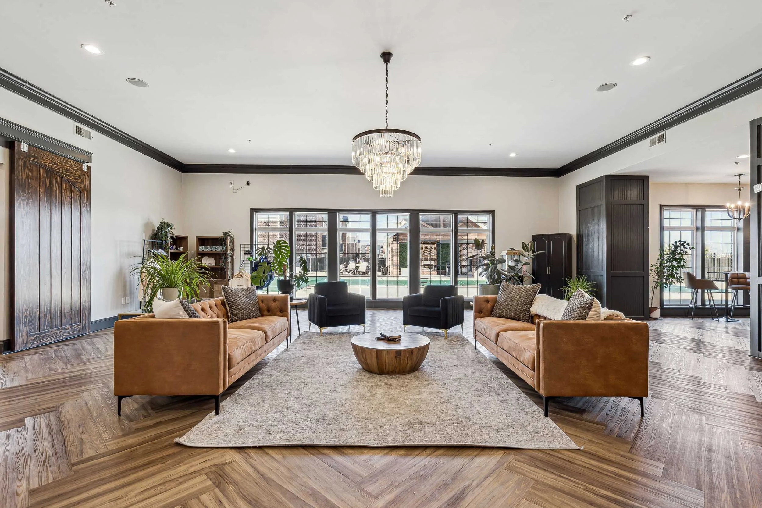 Resident lounge with large windows, six black and tan sofas, a round wooden coffee table, a patterned rug, indoor plants, a crystal chandelier, and a view of a pool outside.