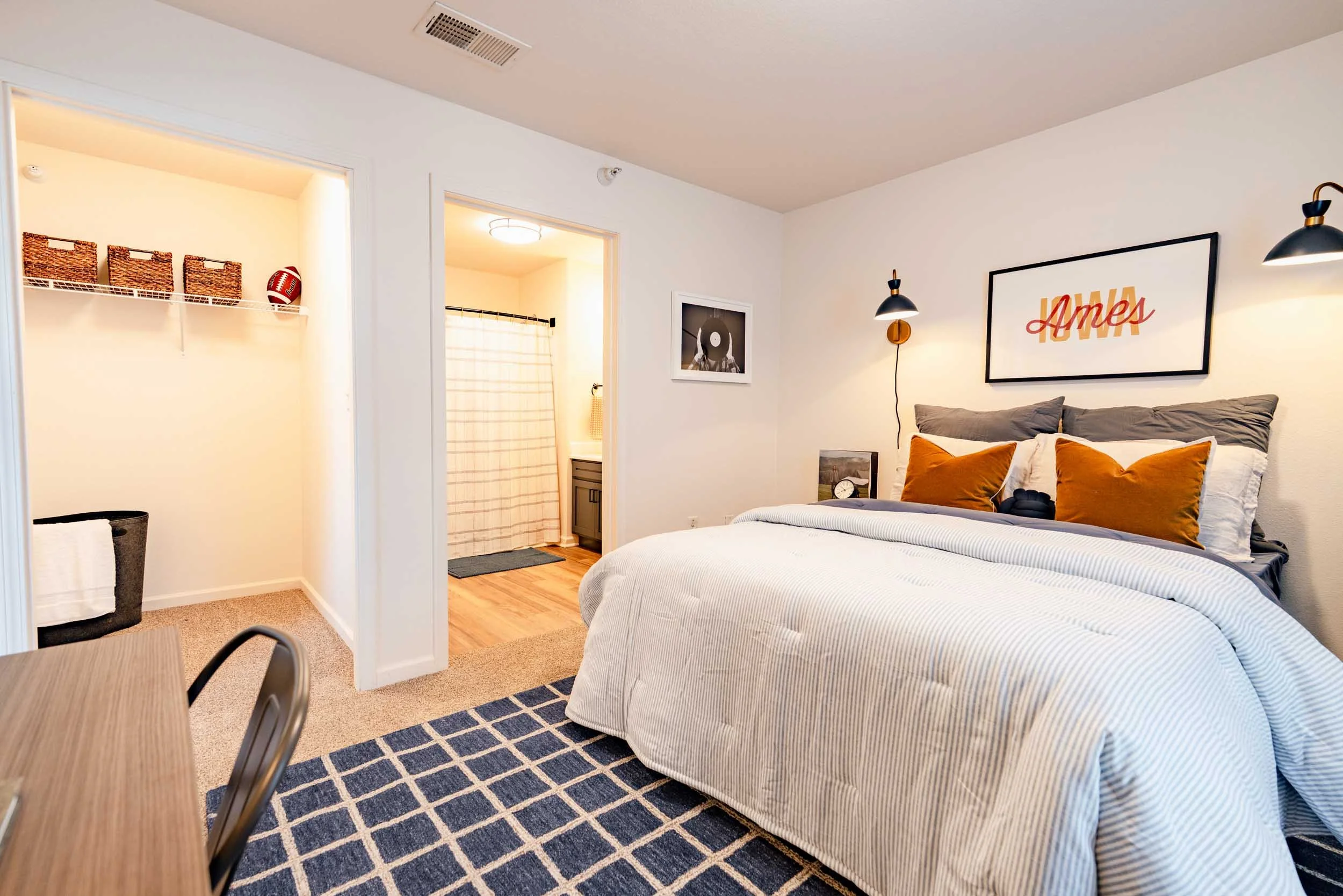 A bedroom with a bed that has gray and orange pillows, a framed 'HOME' art piece on the wall, a bedside clock, and wall-mounted lamps. An open door reveals a bathroom with a shower curtain and a small bathroom vanity.