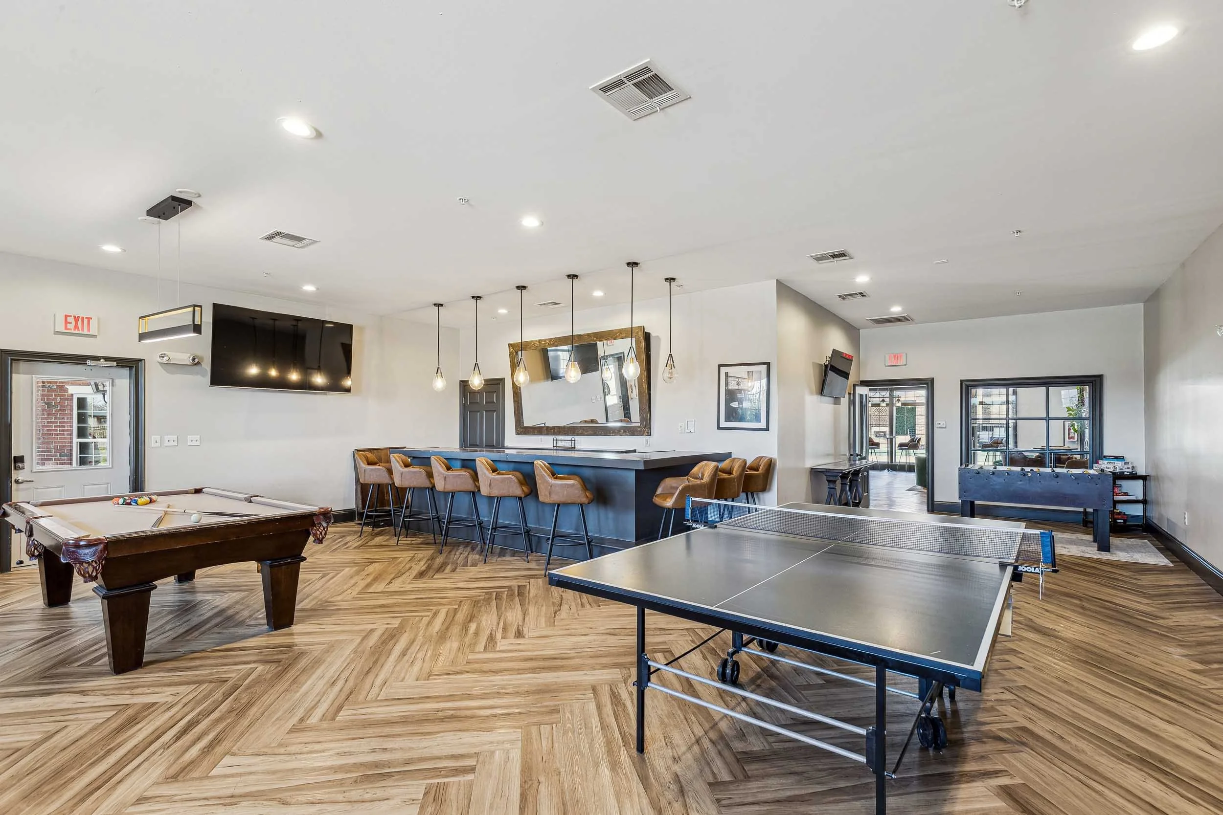 An indoor recreational room with a pool table and a ping pong table, featuring wood flooring, multiple chairs, and hanging lights, with a flat-screen TV and large windows in the background.