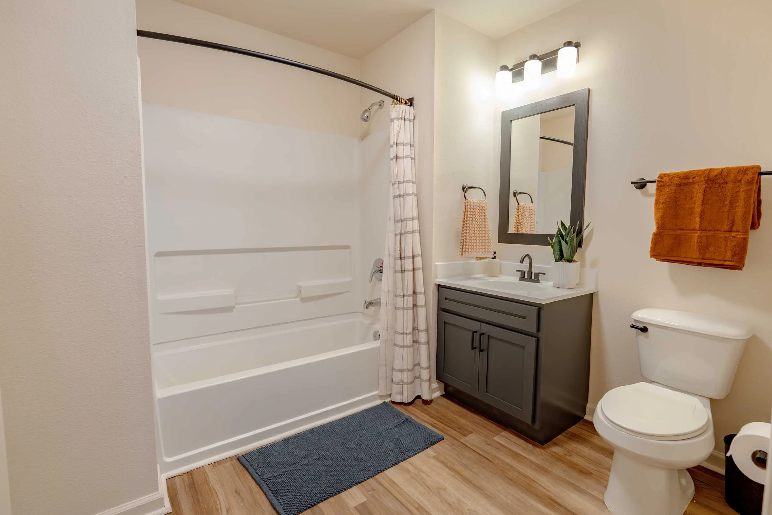 A compact bathroom with a bathtub and shower with a curved shower curtain rod and plaid shower curtain. A dark gray vanity with a single sink, a mirror, and three lights above it. Two orange towels on a black towel rod, a small plant, and a toilet. W