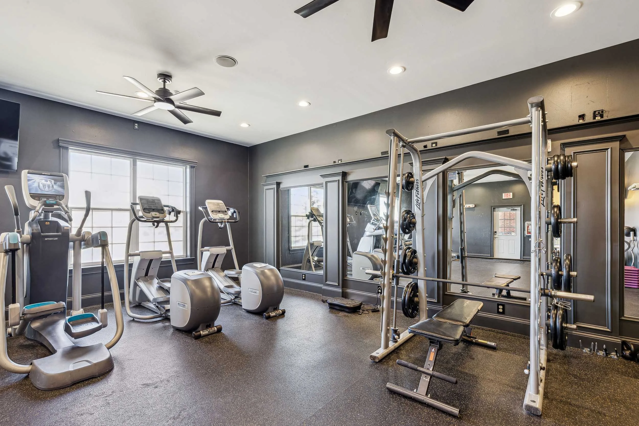 A gym with exercise machines including elliptical trainers, stationary bikes, a weight-lifting station with weights, and a large mirror on the wall. The room has gray walls, large windows, and recessed lighting.