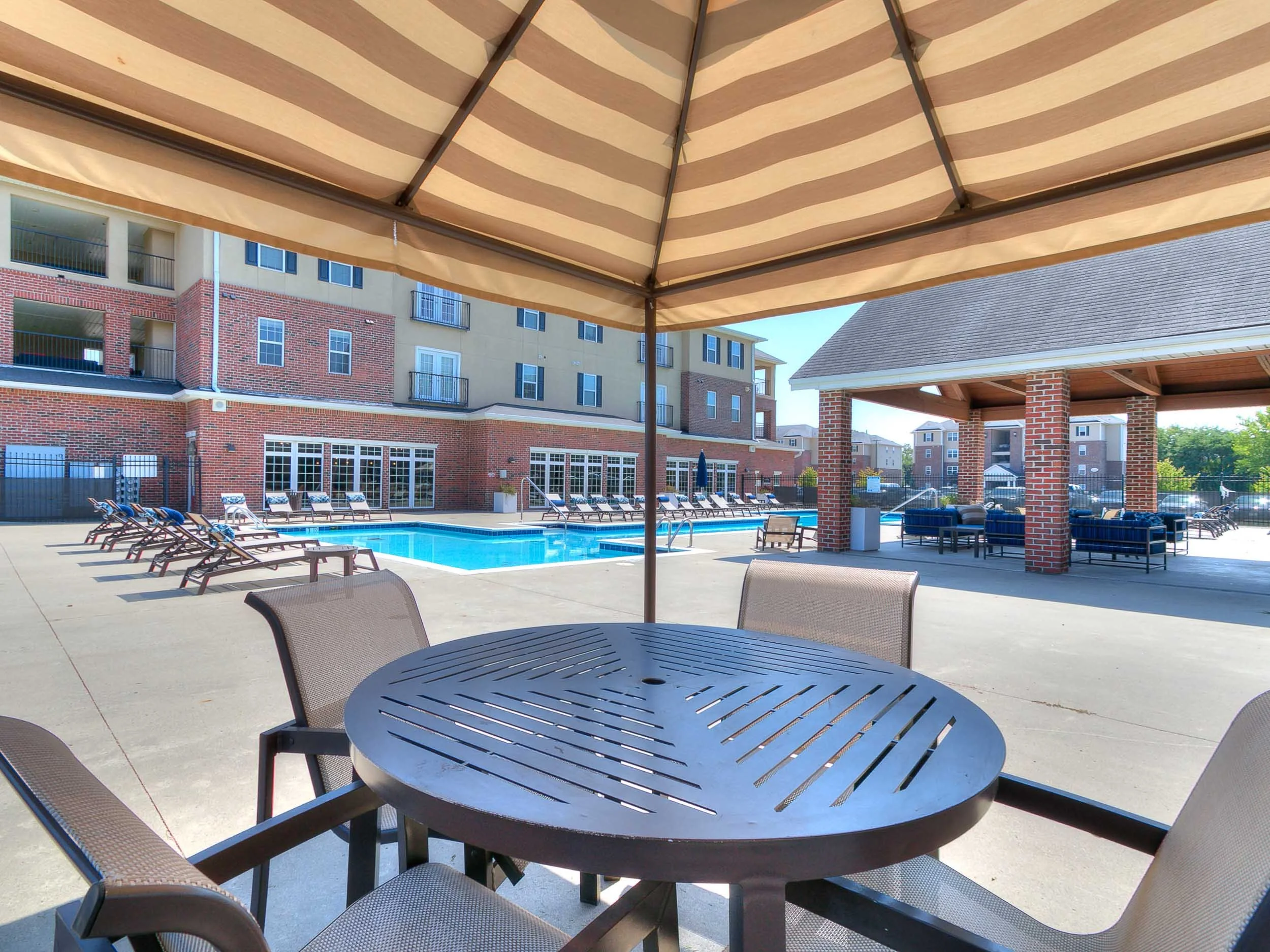 View of an outdoor swimming pool area with lounge chairs, tables, and a shaded pavilion, surrounded by multi-story residential buildings.