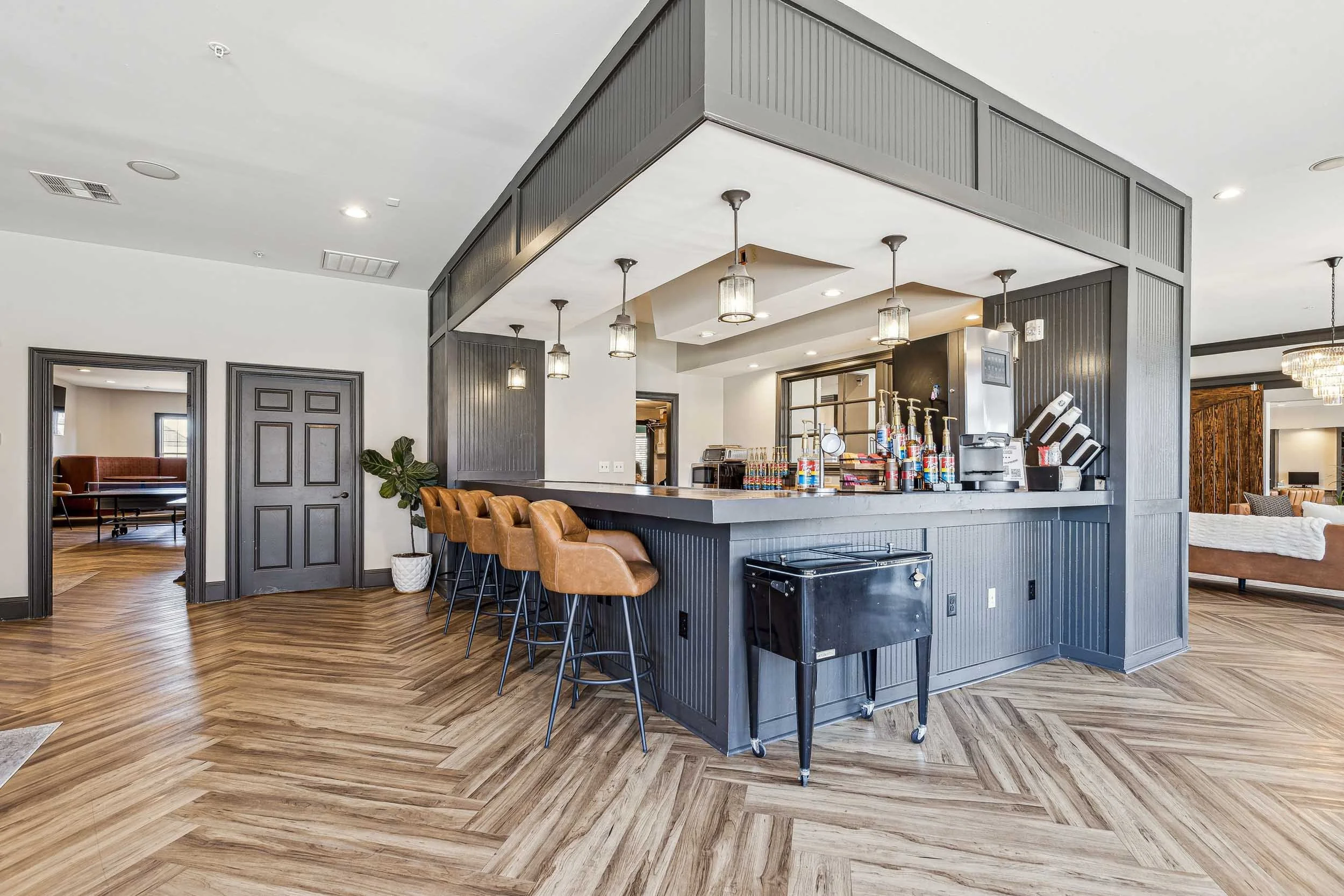 Interior of a modern resident lounge with a bar counter, barstools, hanging pendant lights, and seating areas with brown leather chairs and a sofa. The space has wood flooring and neutral-colored walls.