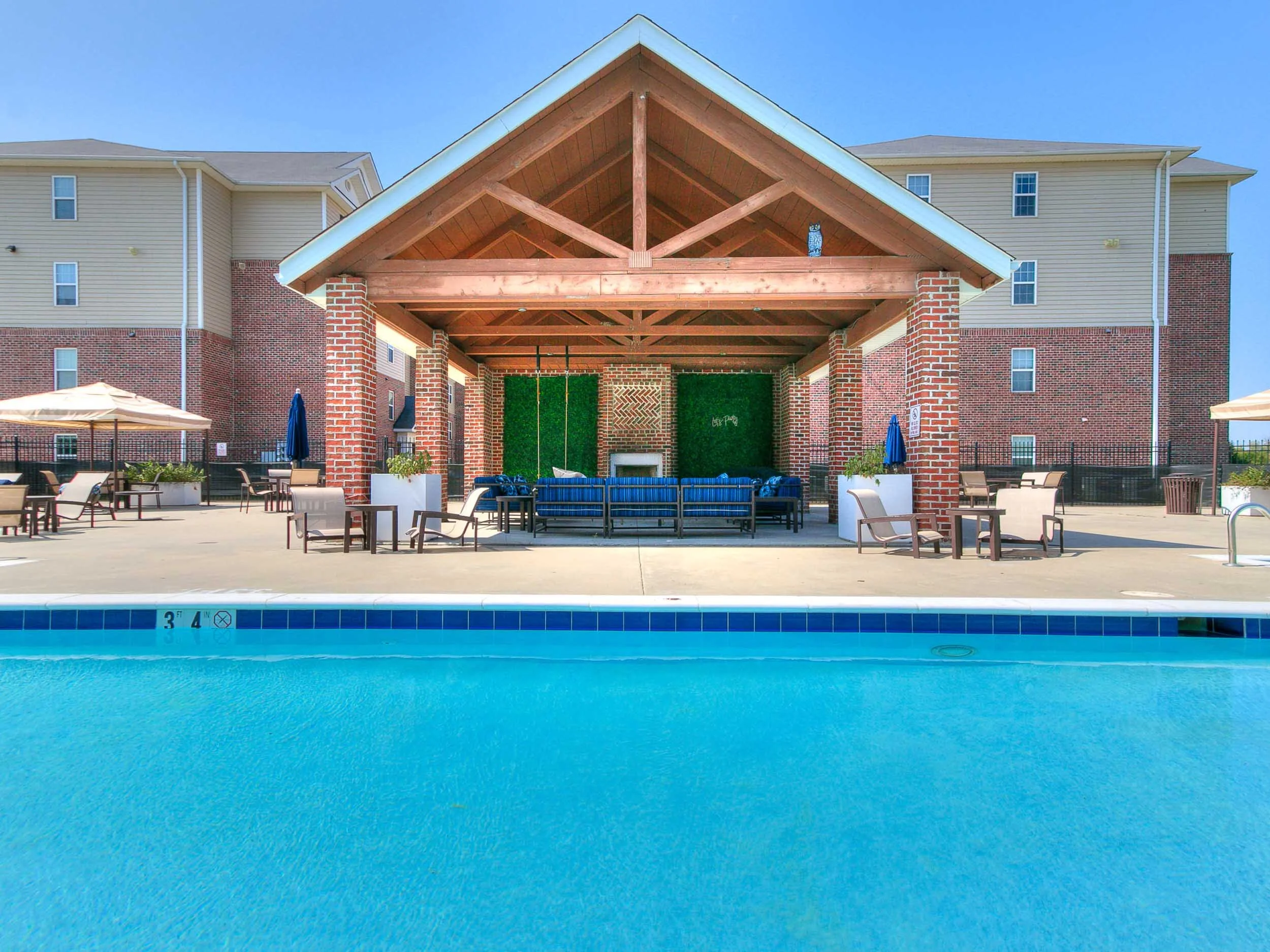 An outdoor swimming pool area with poolside seating and umbrellas, a brick and beige apartment building in the background, and a covered patio with wooden beams and patio furniture.