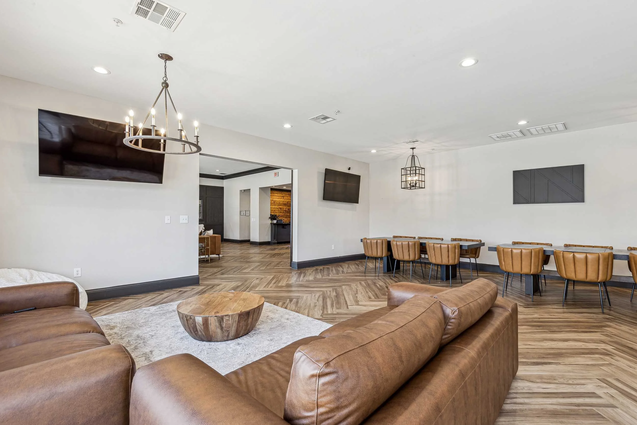 Resident lounge with brown leather sectional sofa, wood coffee table, and dining area with tan chairs. White walls, wooden floor, ceiling lights, two flat-screen TVs, and decorative lighting fixtures.