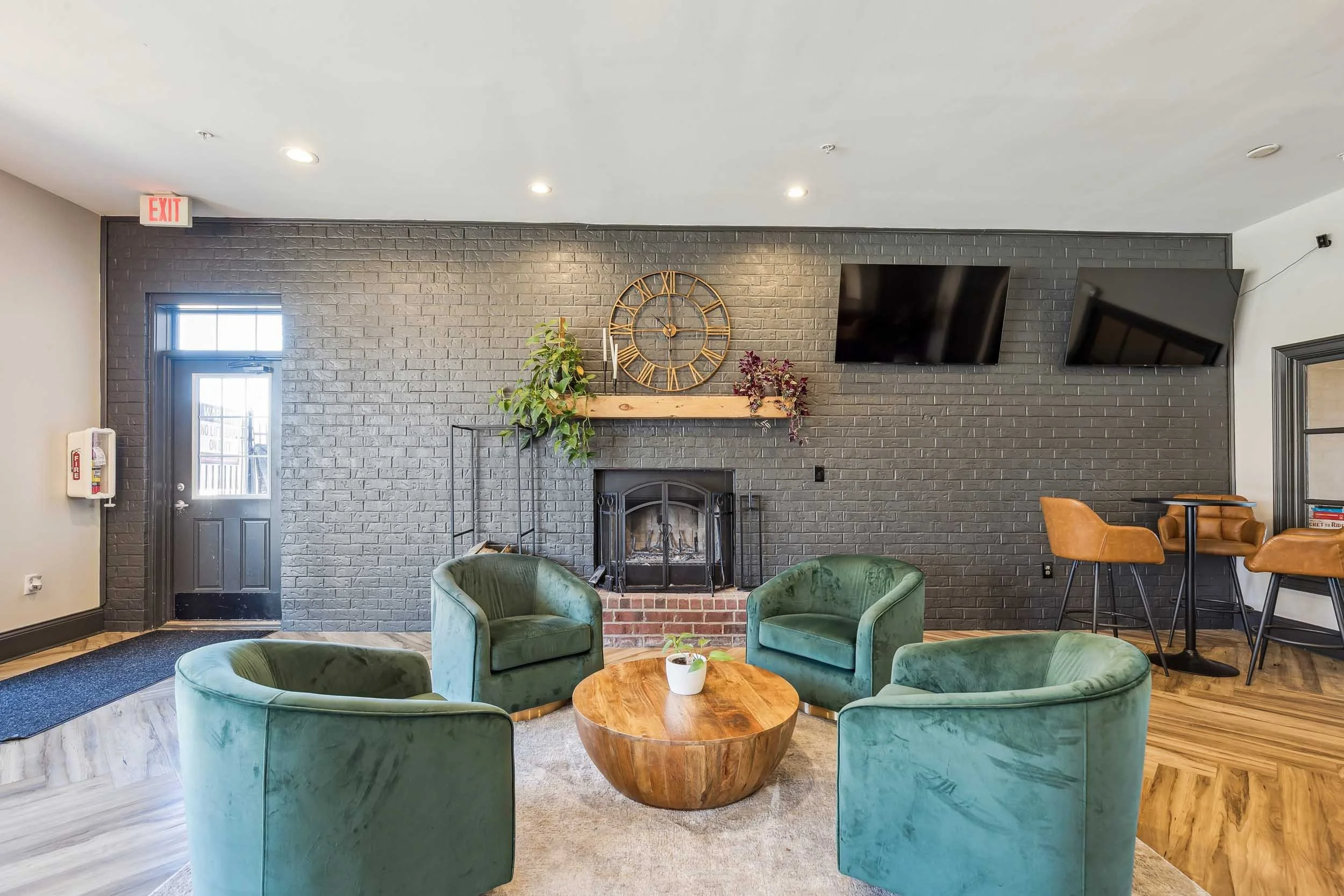 A cozy resident lounge with a black brick fireplace, a round wooden coffee table, green velvet armchairs, a small potted plant, wall-mounted televisions, and decorative plants on a fireplace mantel.