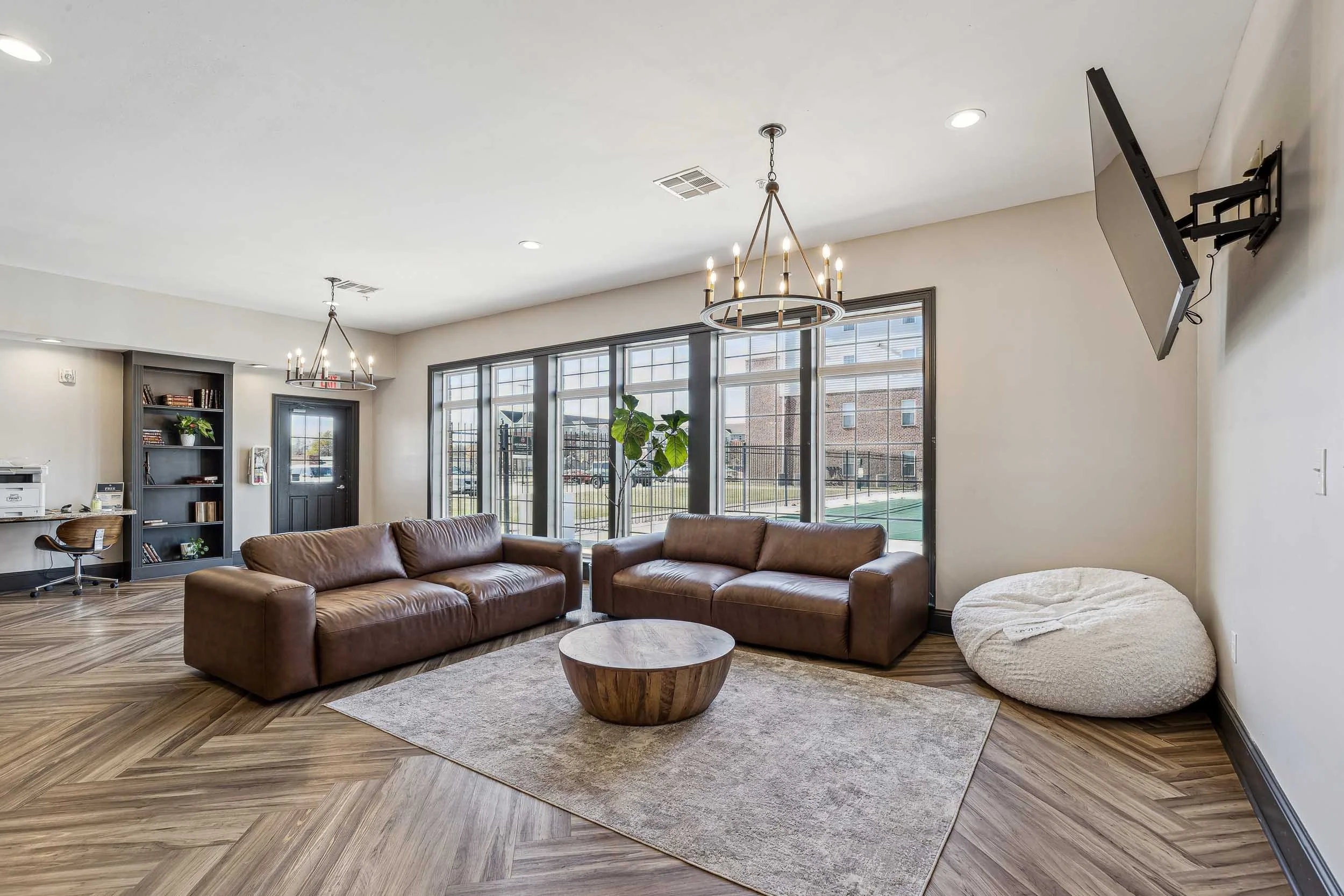 Resident lounge with large windows, leather sofas, a round wooden coffee table, a beige plush pet bed, a wall-mounted TV, and a ceiling chandelier with candles.