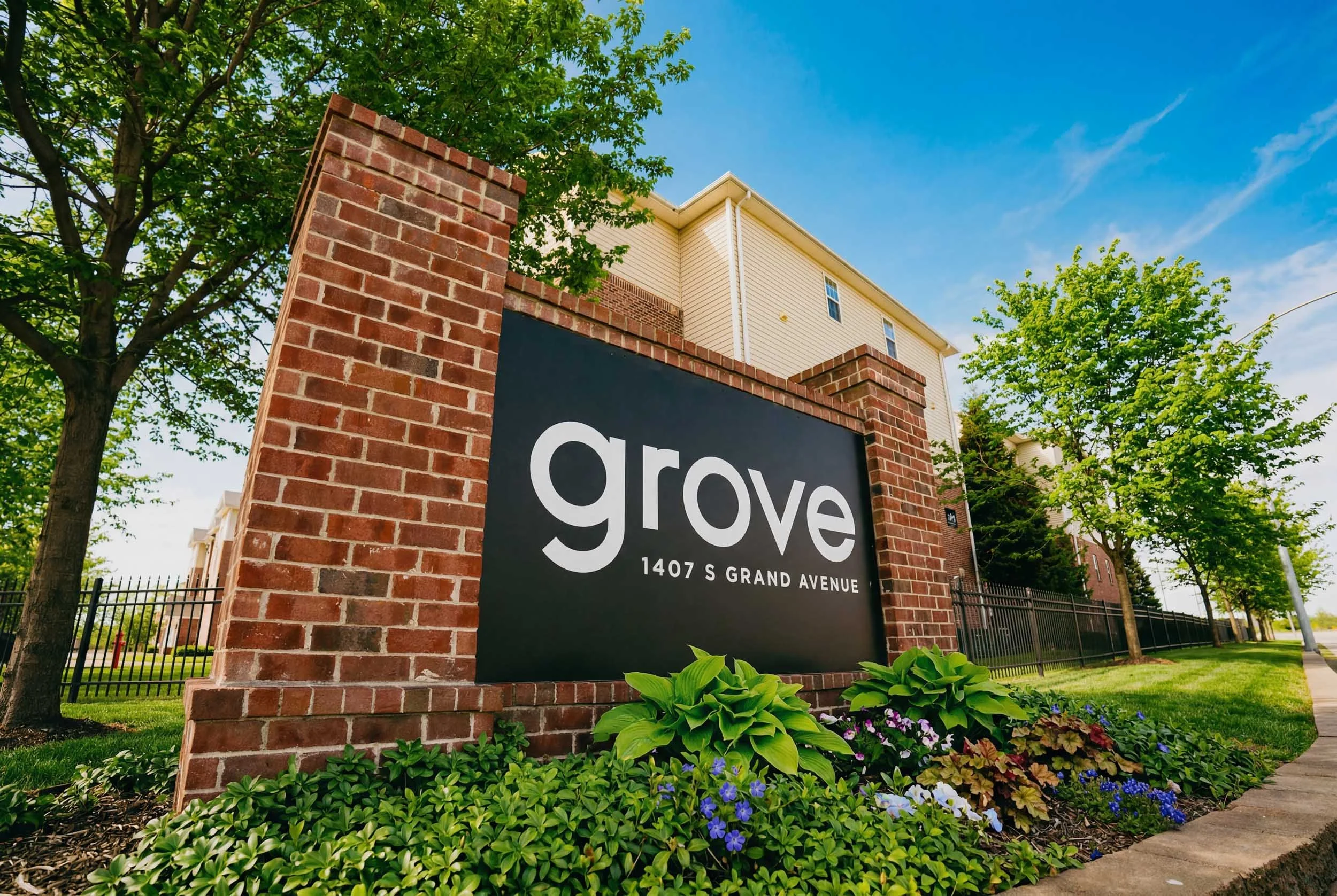 A brick sign with the word 'Grove' and the address 1407 S Grand Avenue, located in front of a landscaped area with green plants and colorful flowers. In the background, a beige residential building with trees and a blue sky with some clouds.