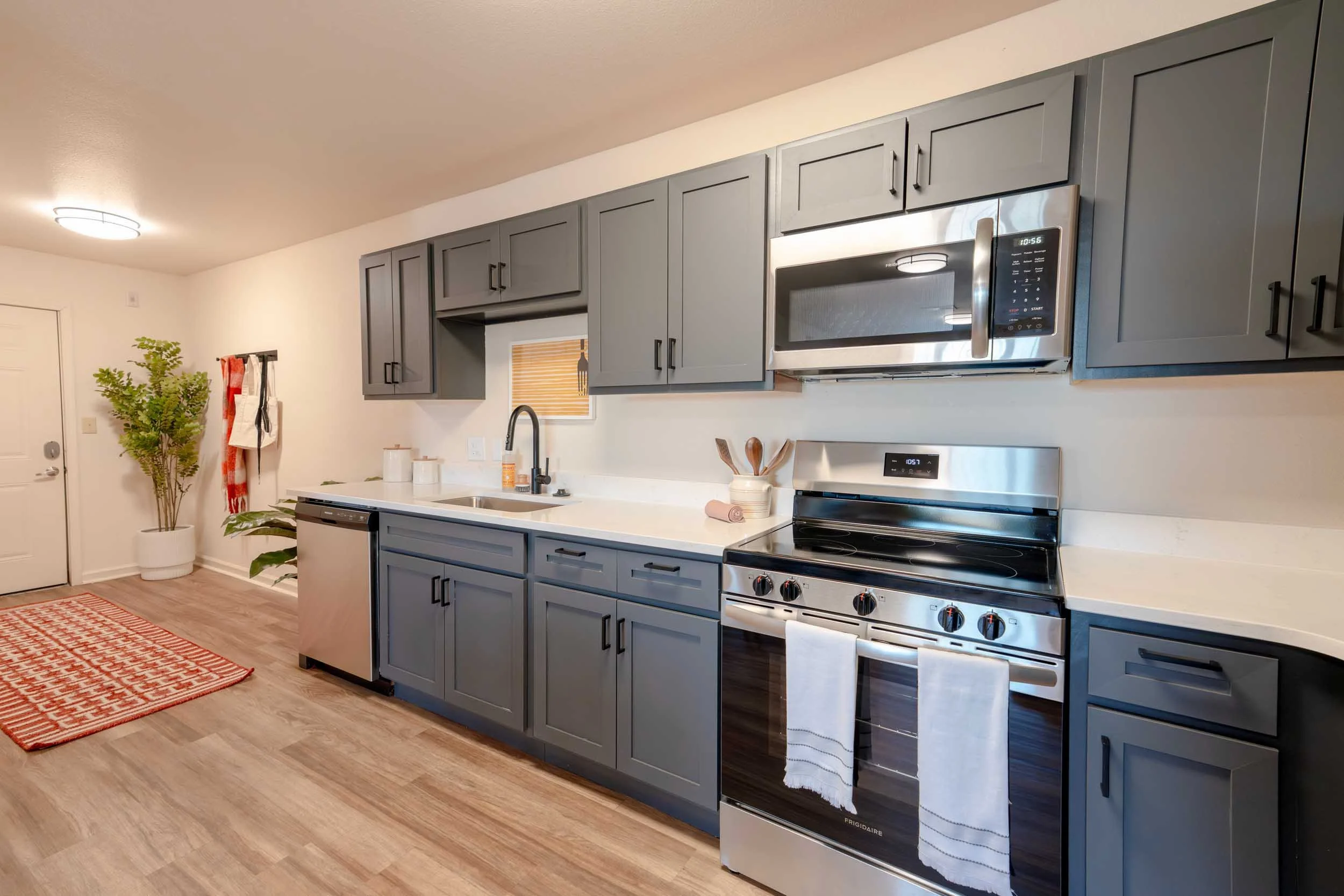 Modern kitchen with gray cabinets, stainless steel microwave and oven, black stovetop, white countertop, plants, and a red patterned rug on wood floor.