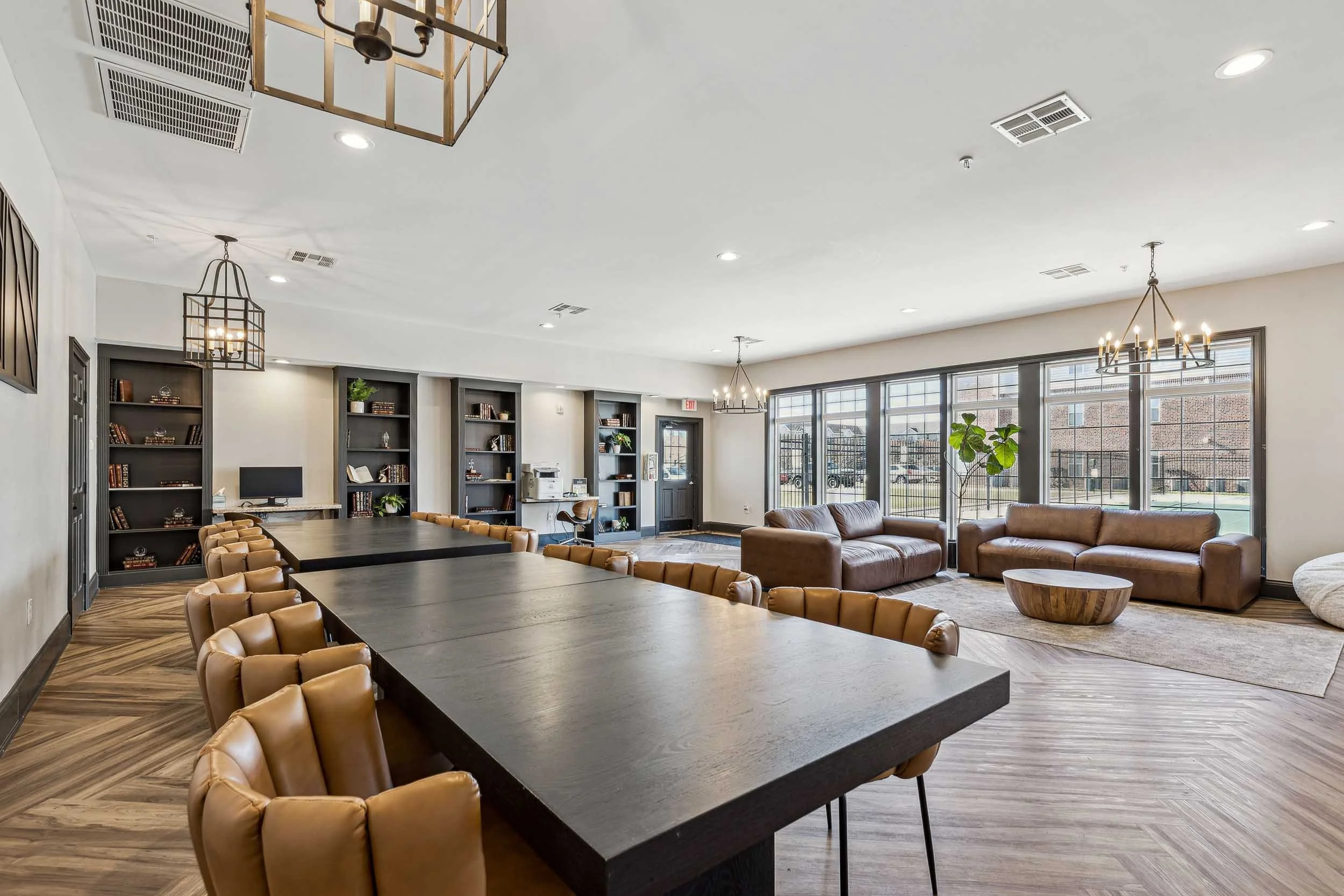 A spacious, modern lounge area with large windows, beige leather armchairs, a dark coffee table, and wooden flooring. There are bookshelves, ceiling lights, and decorative chandeliers.