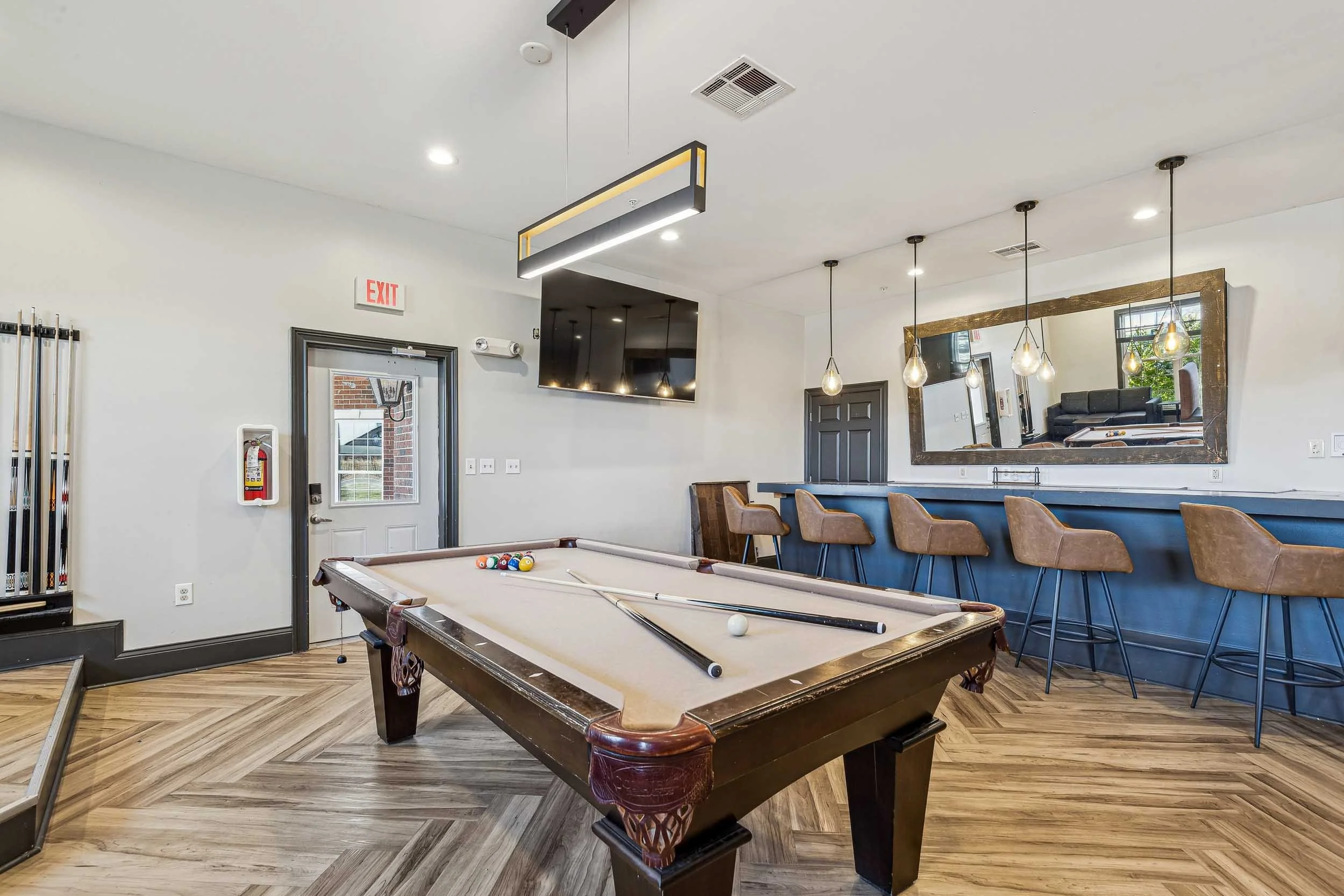 Indoor recreation room with a pool table, pool balls, and cues, a large mirror, a TV, and bar seating with wooden chairs, modern light fixtures, and wood flooring.