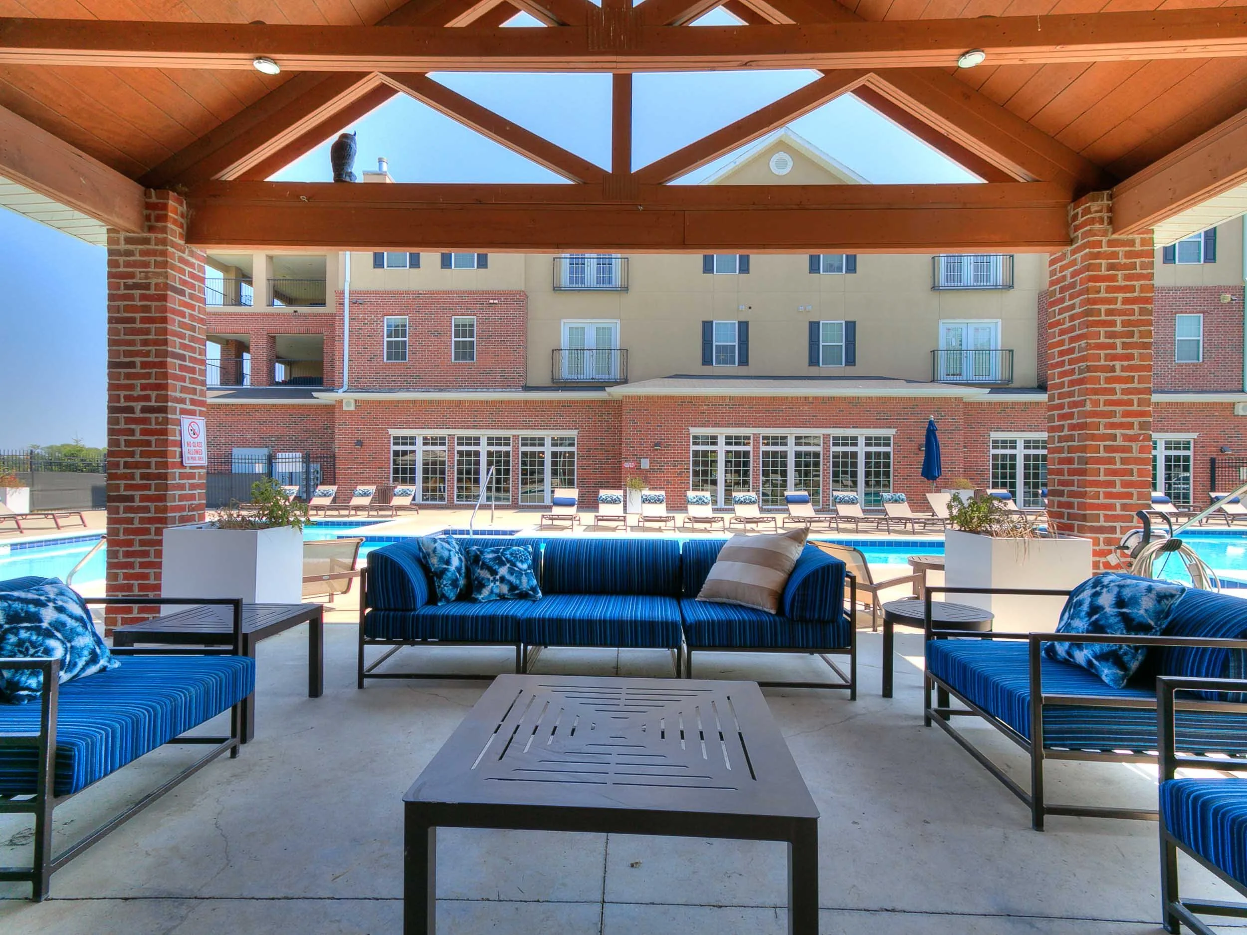View from beneath a wooden gazebo with blue and white striped outdoor furniture around a pool, apartment building with balconies in the background, and a blue sky.