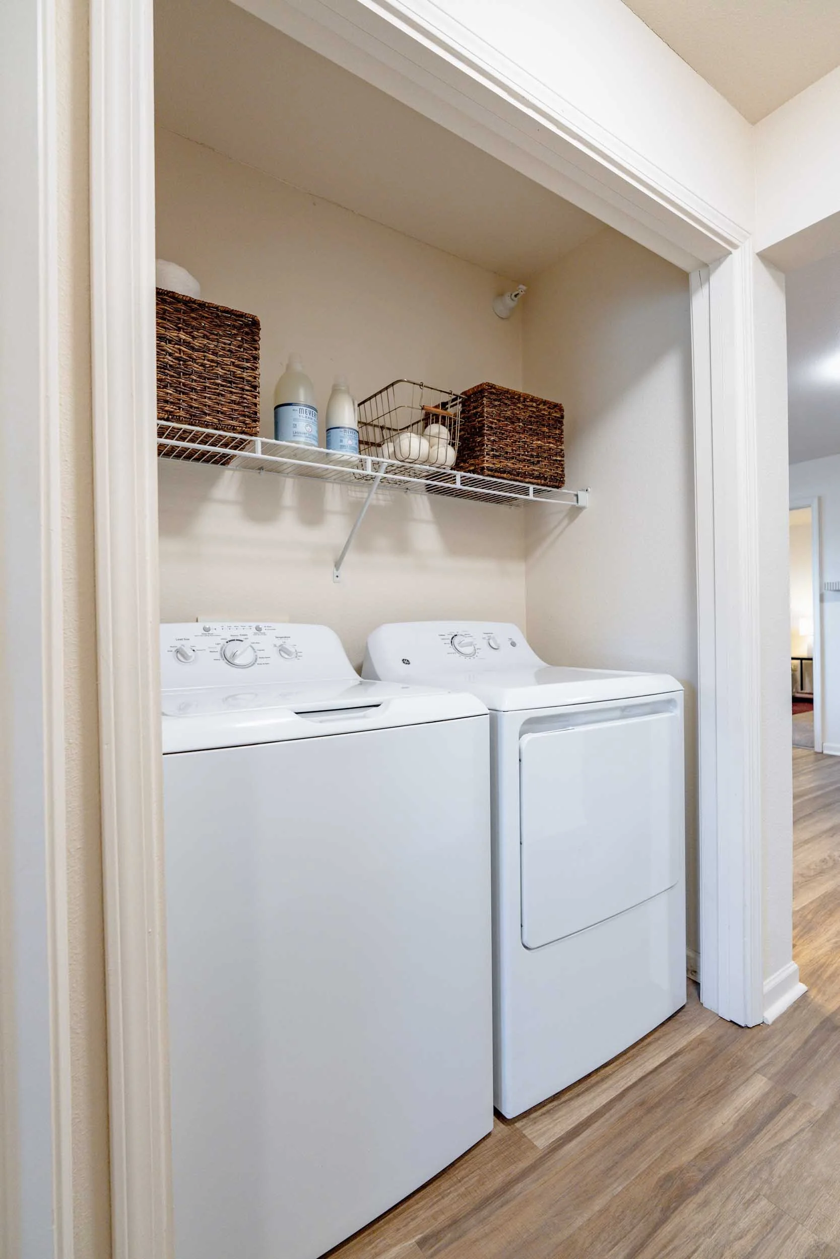 Front view of a laundry room featuring a top-loading washing machine and a front-loading dryer, with a wood shelf above containing laundry supplies and storage baskets.