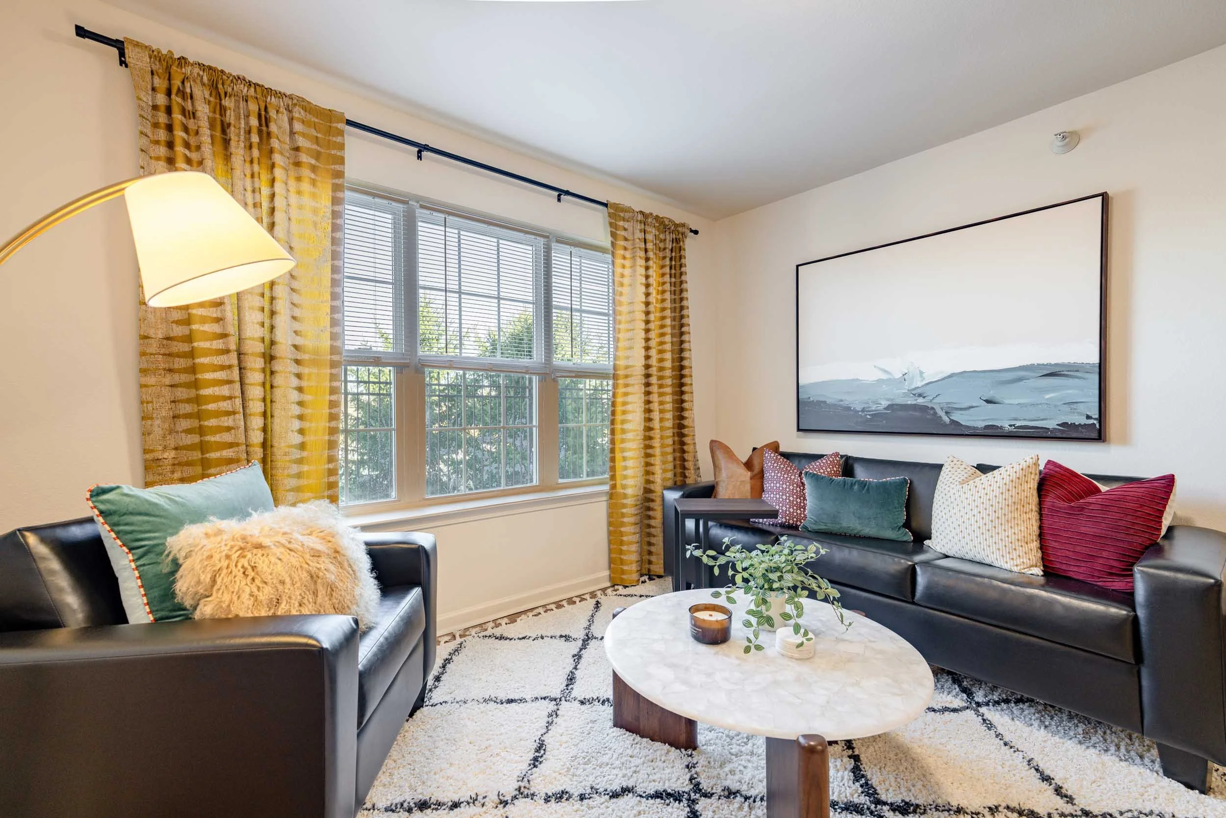Living room with black leather sofas, colorful pillows, a round marble coffee table, a window with yellow curtains, wall art, and a lamp.