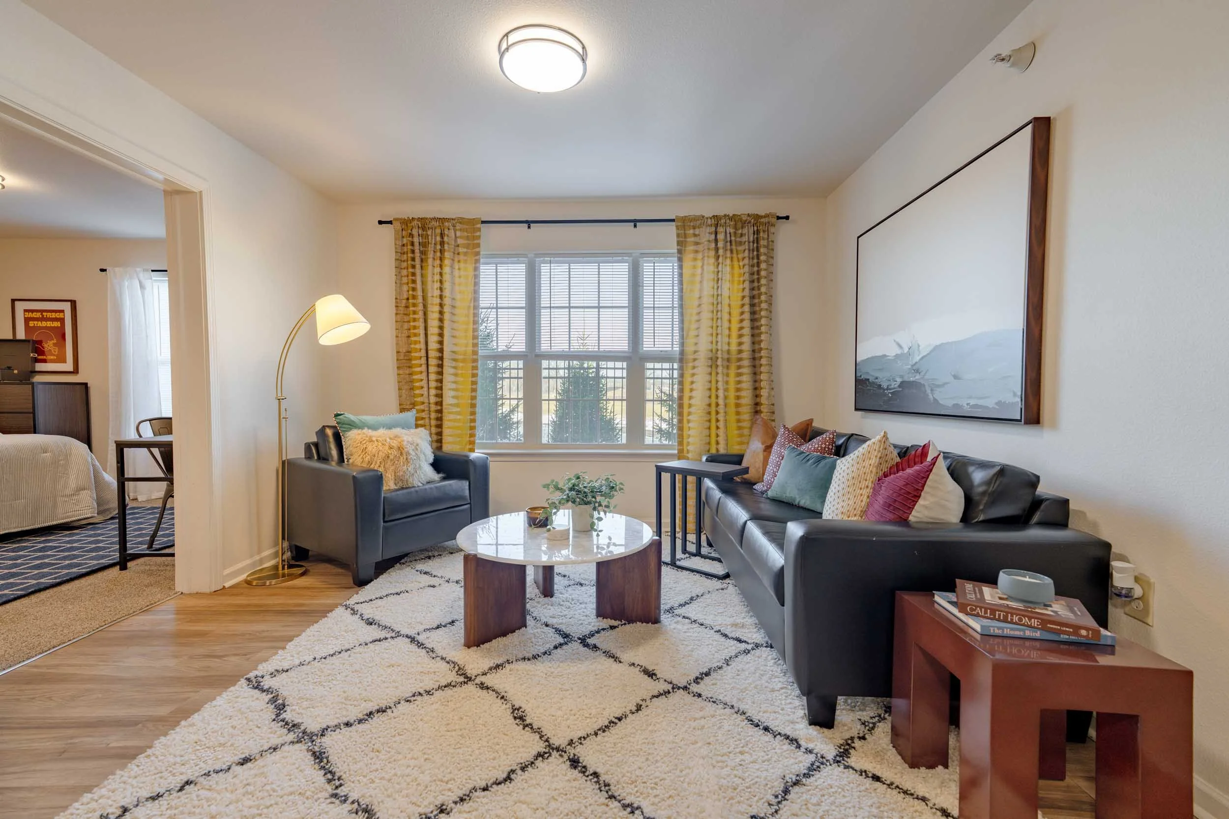 A living room with a window, yellow curtains, modern black leather sofas with colorful pillows, a round coffee table, a side table with books, a floor lamp, and artwork on the wall.