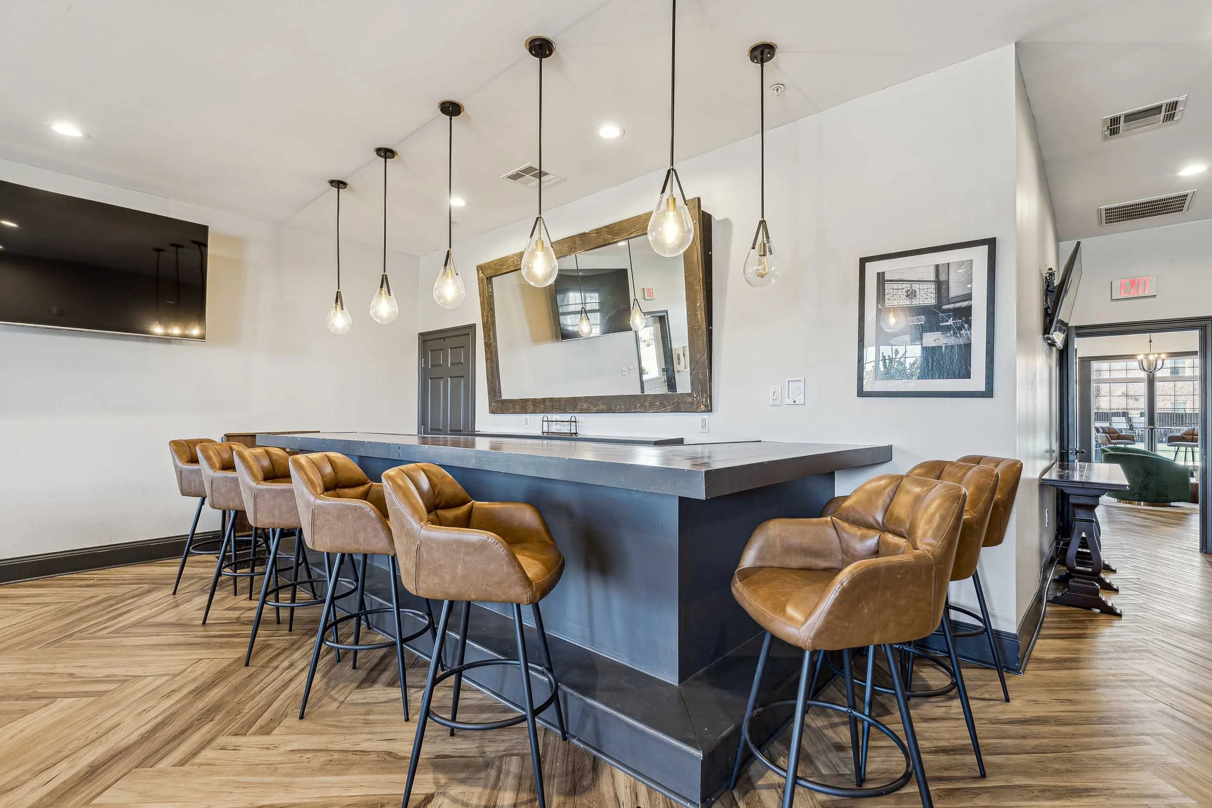 Modern interior with a bar counter and brown leather chairs around it, pendant lights hanging from the ceiling, large mirror on the wall, television, framed picture, and a doorway leading to another room.