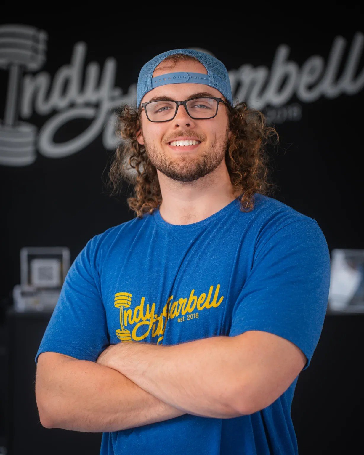 A man with curly hair, wearing glasses, a blue baseball cap backwards, and a blue T-shirt with yellow text, standing indoors with arms crossed, smiling at the camera.