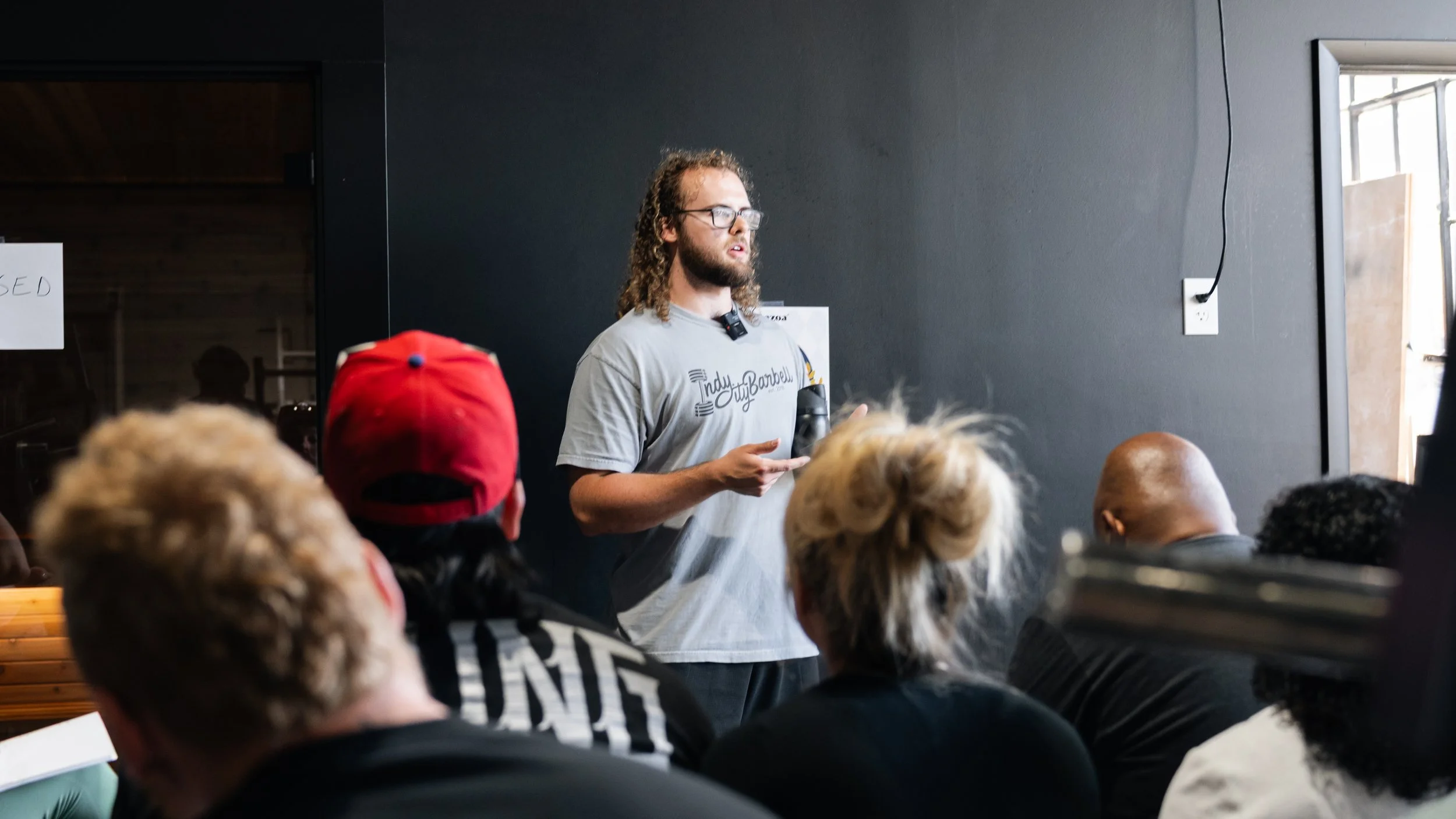 A man with long curly hair, glasses, and a beard speaking in front of a small audience in a room. He is wearing a gray T-shirt with a logo and holding a black object, possibly a microphone. The audience includes a person in a red cap and others with various hairstyles, sitting and facing him.