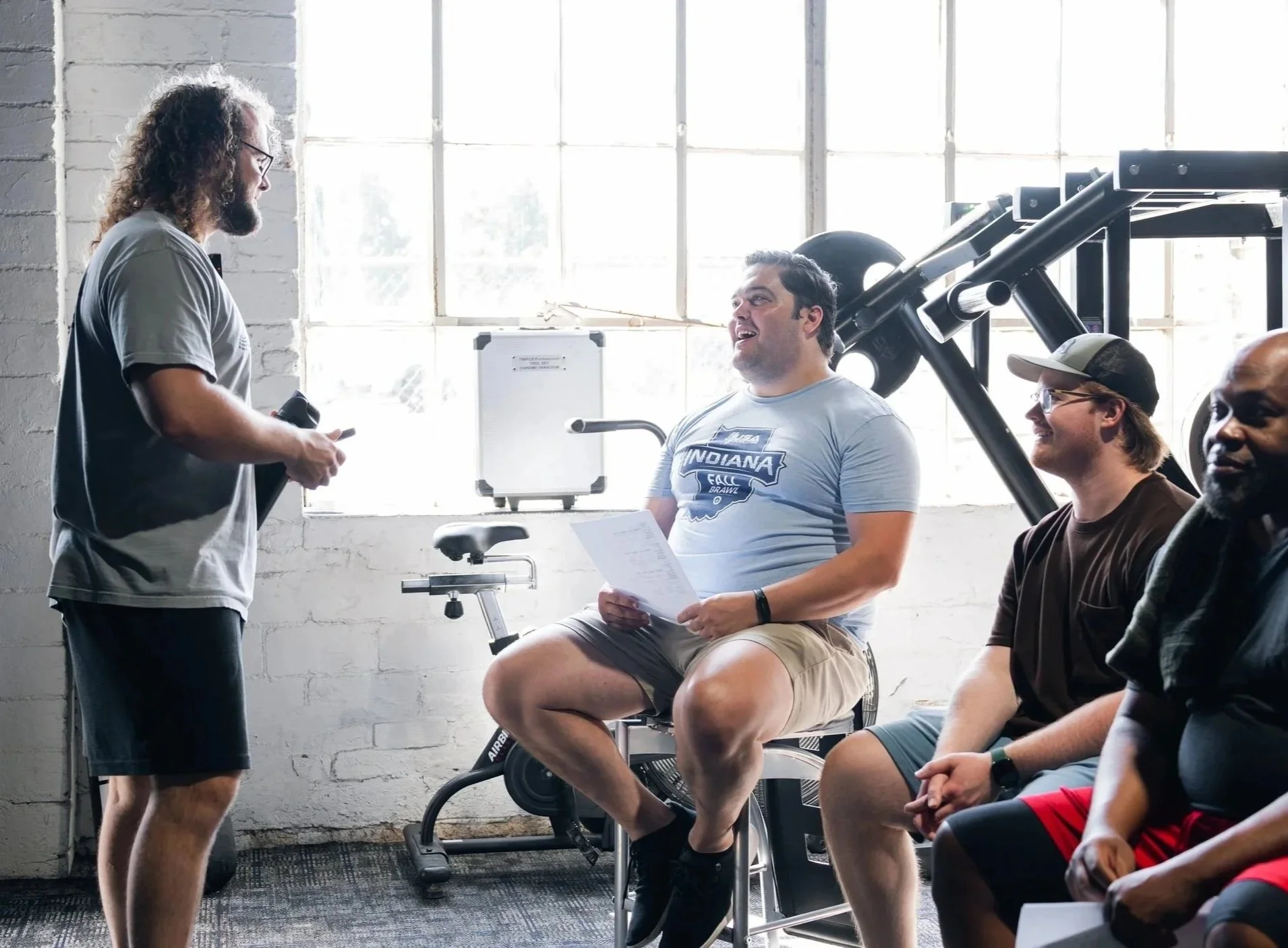 Group of four men in a gym, one standing and three sitting, having a conversation during a workout session. The gym has large windows and exercise equipment.