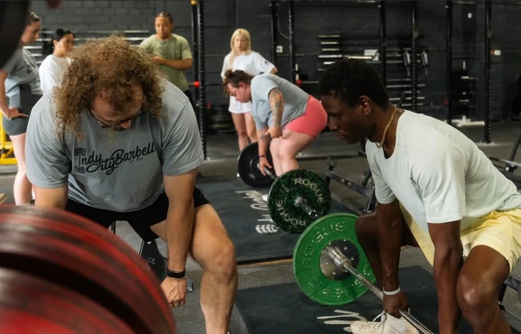 People lifting weights in a gym, with two men in the foreground preparing for deadlifts and others in the background watching.