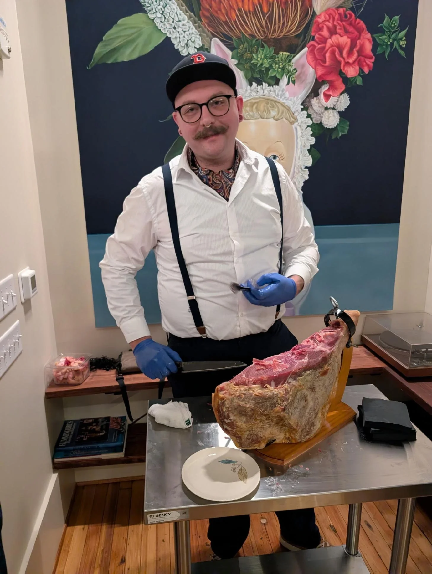 Man with glasses, mustache, and suspenders standing behind a table with a large piece of raw meat and a plate, holding a carving knife and a fork, in front of a colorful floral mural.