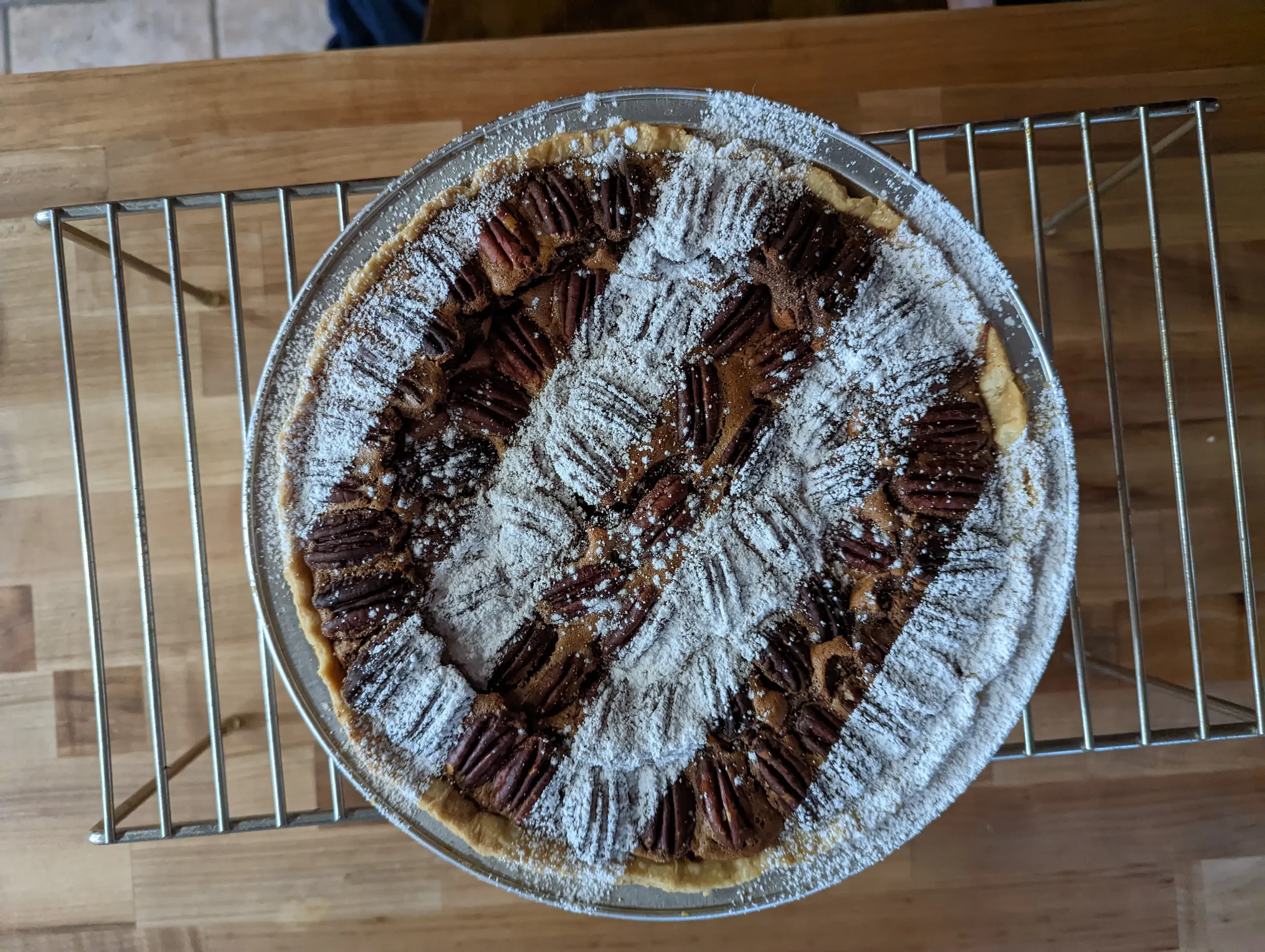 A pecan pie dusted with powdered sugar on top, resting on a wire rack over a wooden surface.