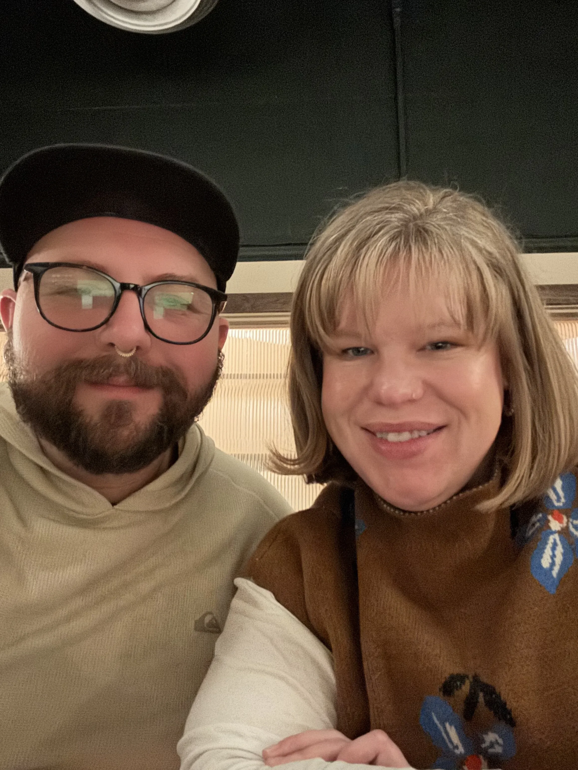 A man with glasses and a black hat, and a woman with shoulder-length blonde hair, both smiling, taking a selfie in an indoor setting.
