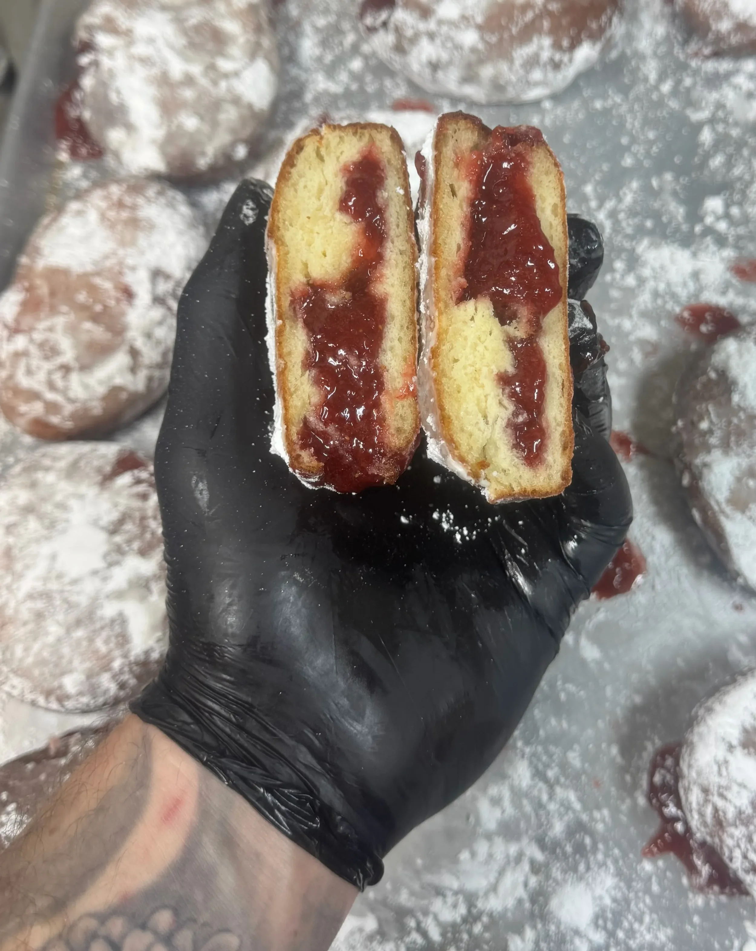 A hand wearing a black glove holding a sliced Twinkie with cream filling and red jam inside, with powdered sugar and additional Twinkies in the background.