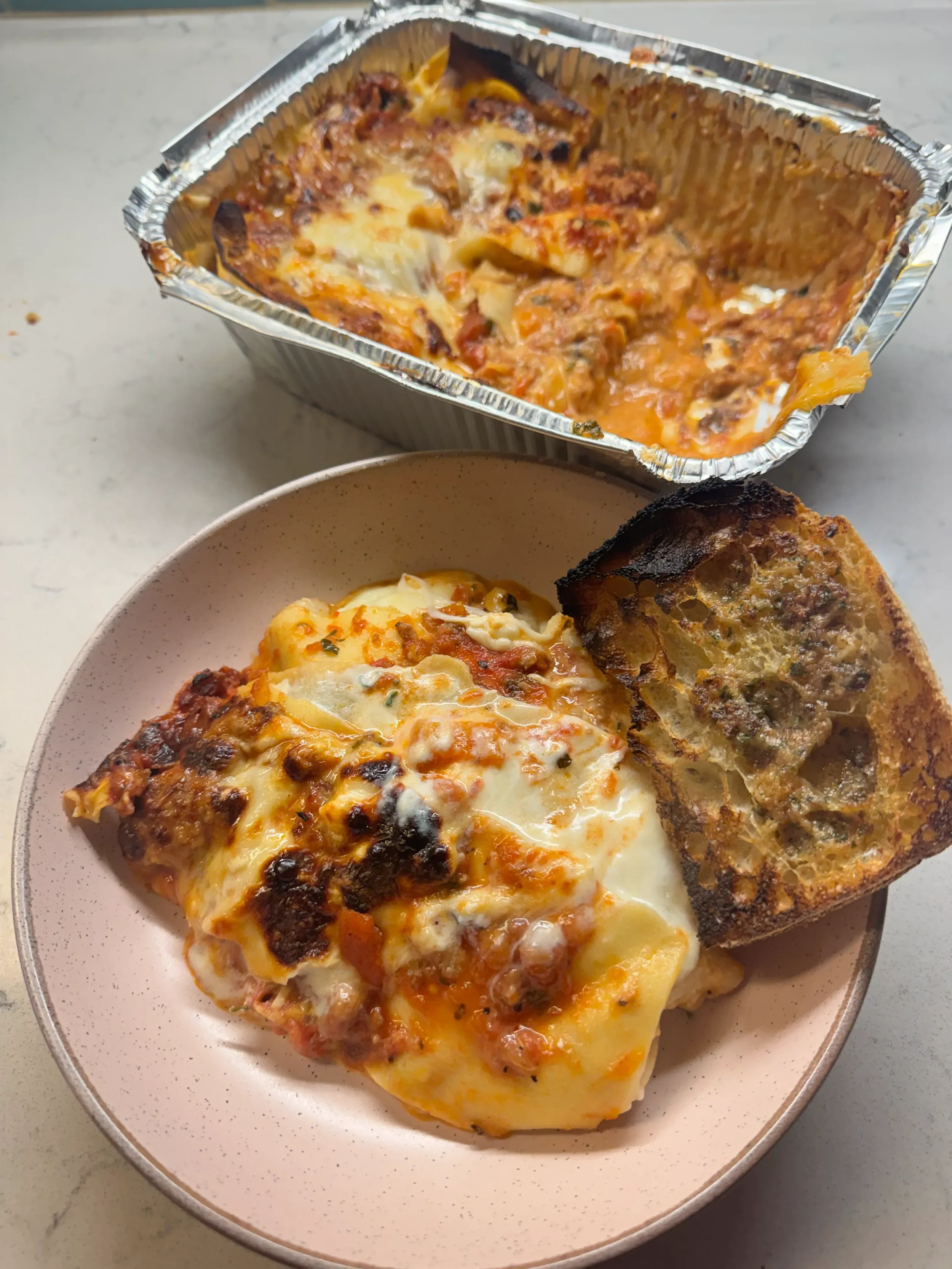 A plate containing a serving of baked lasagna with melted cheese and a slice of toasted garlic bread. In the background, there is an aluminum tray with remaining lasagna.