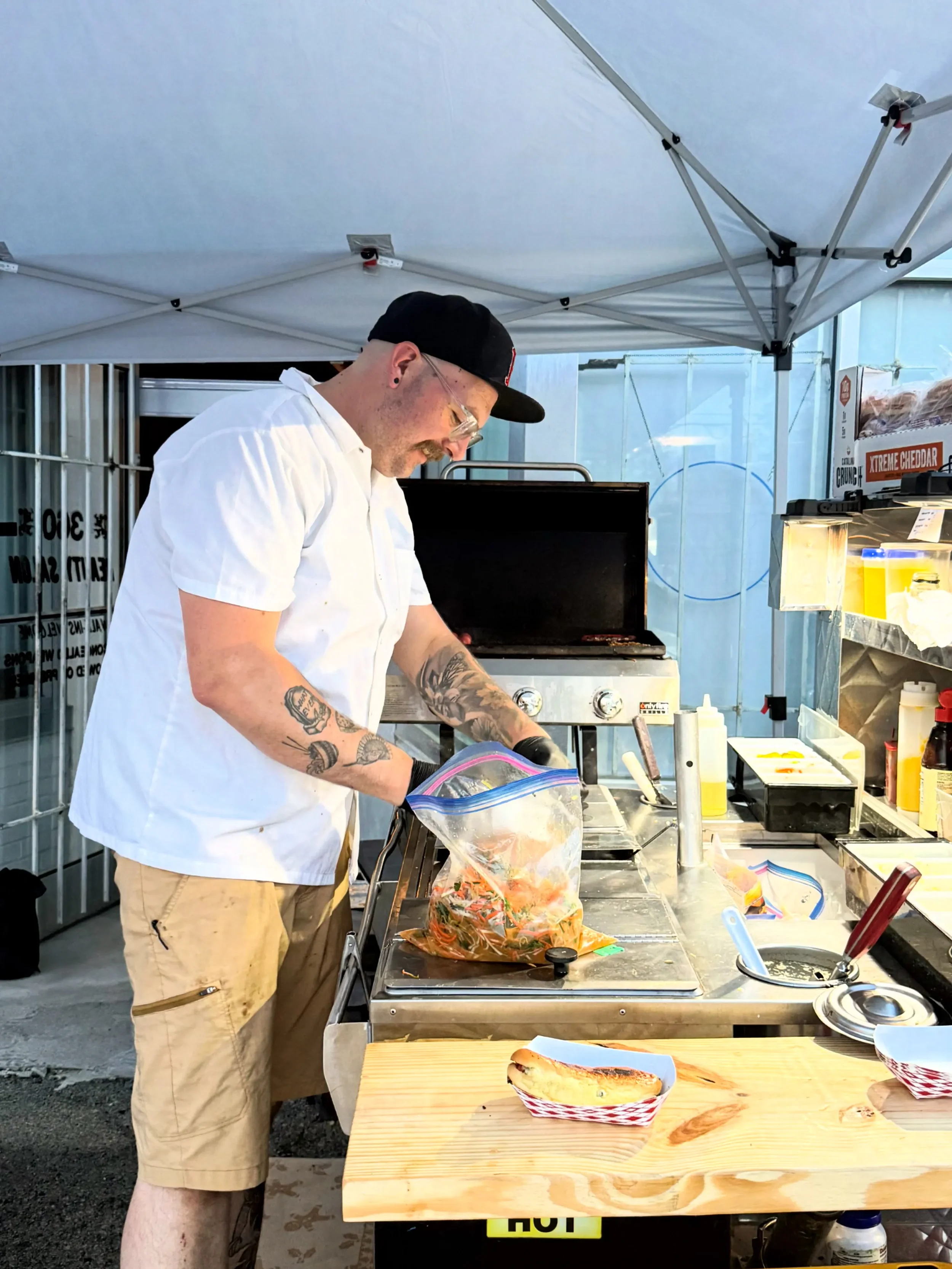 A man with tattoos on his arms, wearing a white shirt, tan shorts, a black cap, and glasses, preparing food at an outdoor food stand under a white canopy. There is a grill, condiments, and a piece of grilled bread on a wooden table in front of him.