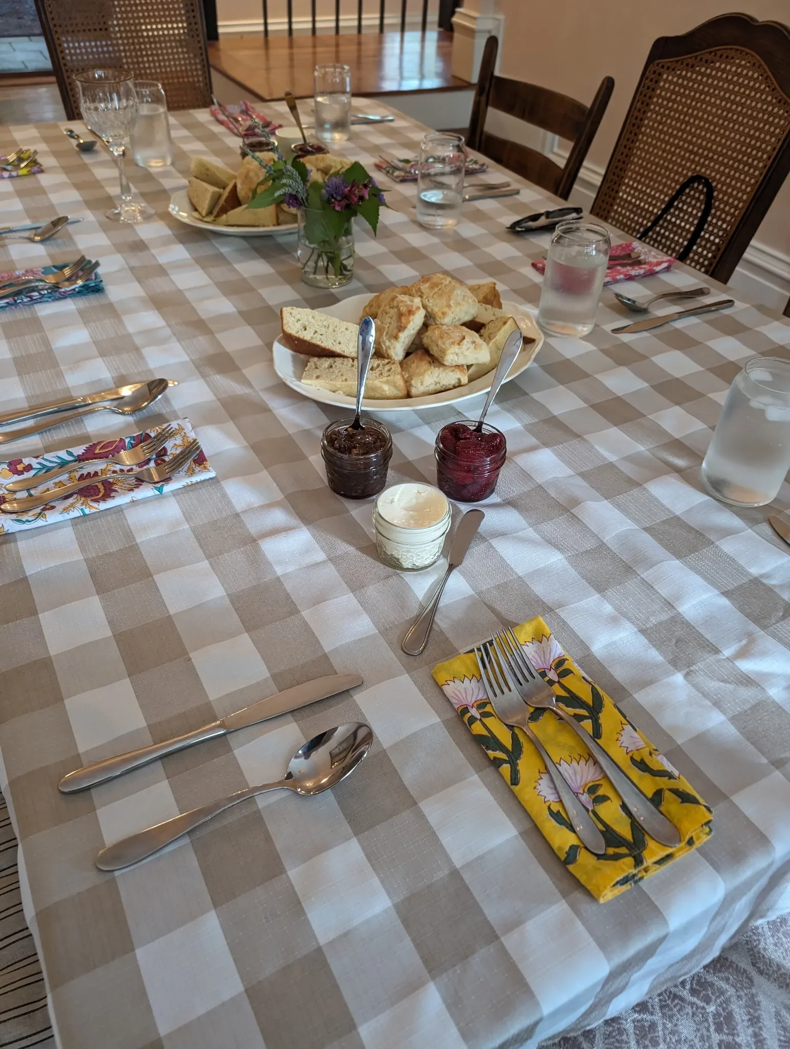 A dining table set for a meal with a checkered beige and white tablecloth, plates of bread, jars of jam, and utensils. There are glasses of water, a small vase with flowers, and colorful napkins.