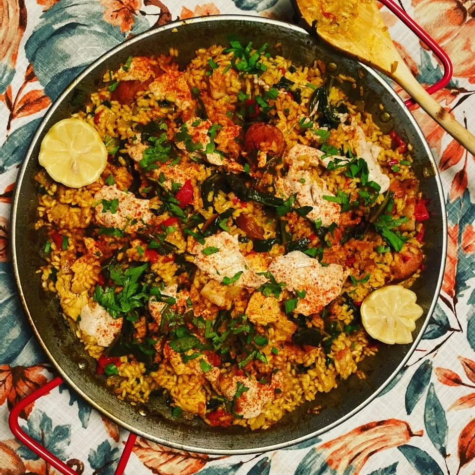A pan of seasoned rice with vegetables, herbs, and chunks of seafood, garnished with parsley and lemon slices, on a floral tablecloth.