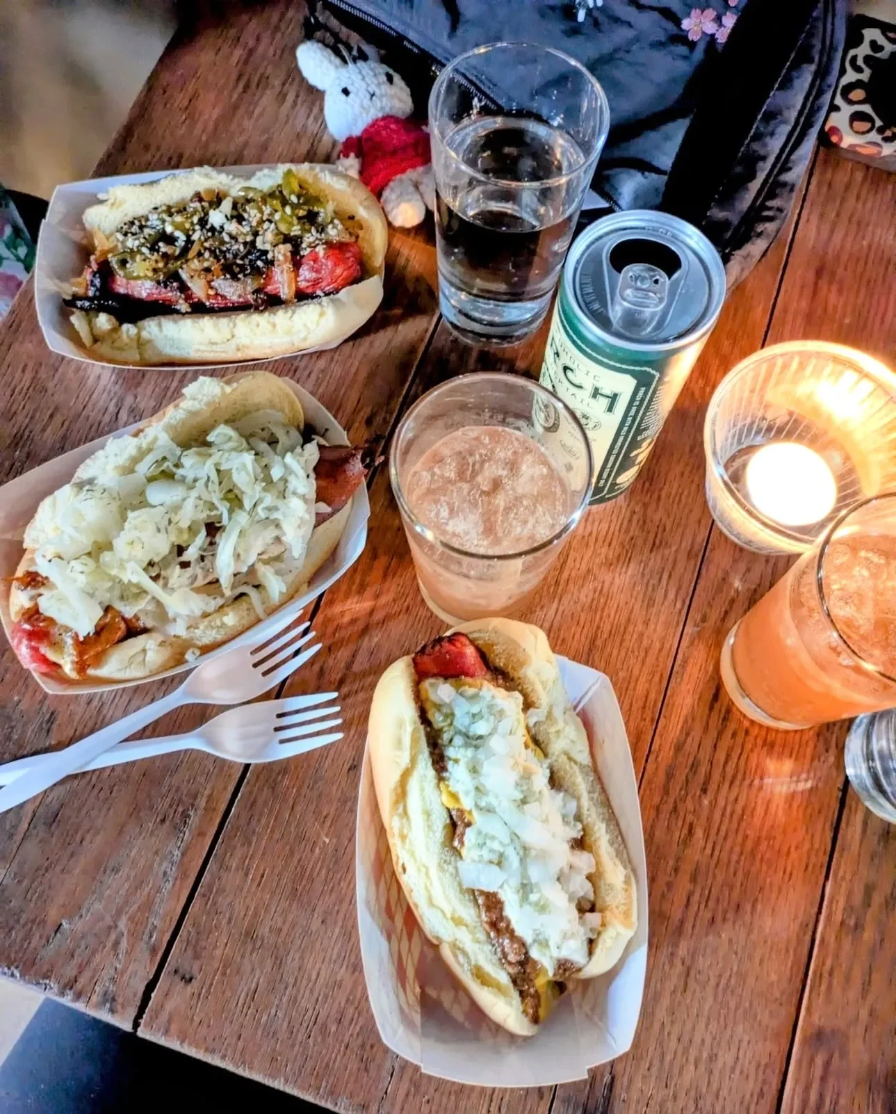 A wooden table with two hot dogs topped with mustard, sauerkraut, and relish, two glasses of water, a can of LaCroix sparkling water, two lit candles, and two drinks in glasses, one possibly orange juice or a cocktail. There is a small plush toy resembling a dog in the background.